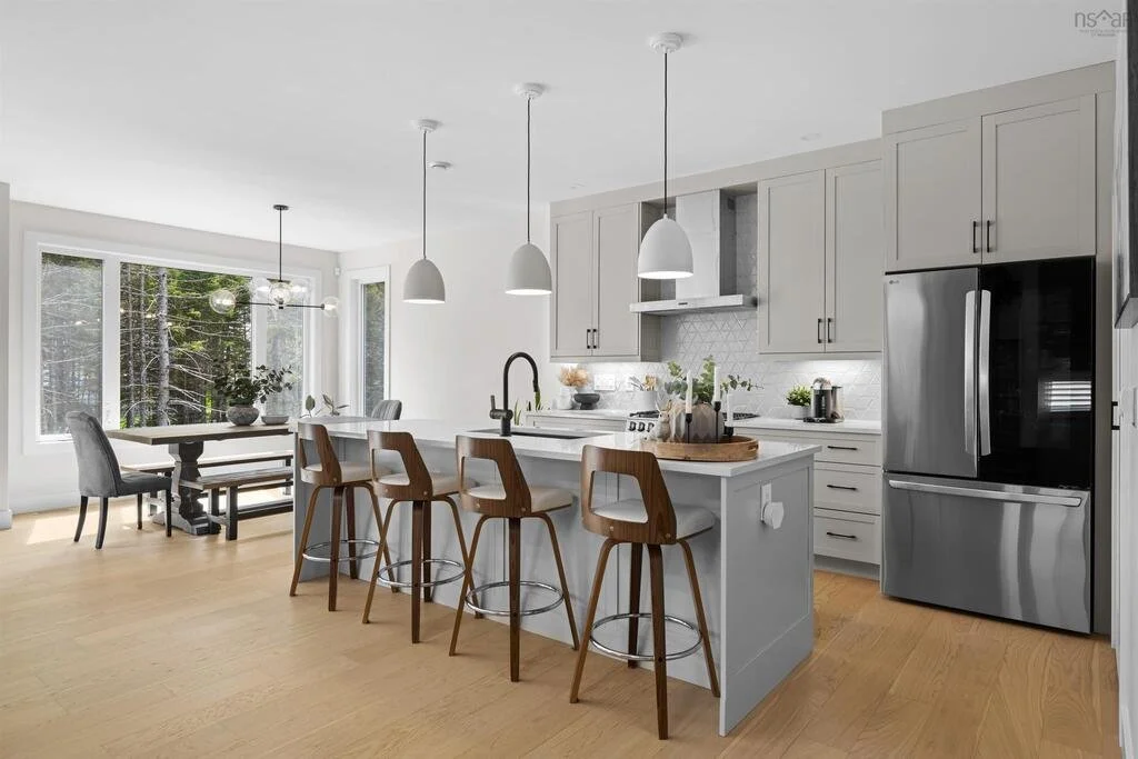Modern kitchen and dining area with large windows, white cabinets, stainless steel refrigerator, island with three wooden bar stools, and pendant lighting.