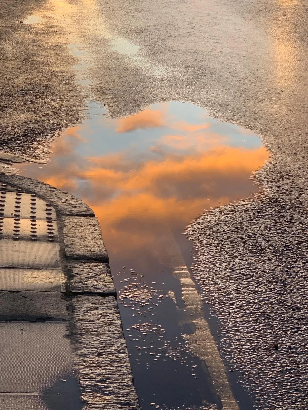 Reflection of a sunset sky with orange clouds in a puddle on the street, with a sidewalk and curb in the foreground.