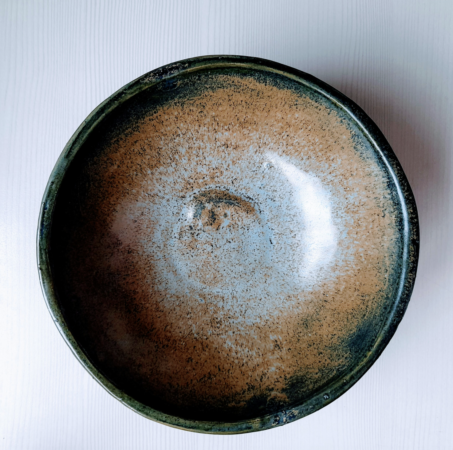 A close-up of a round ceramic or metal dish with a speckled brown and black glaze on a white surface.