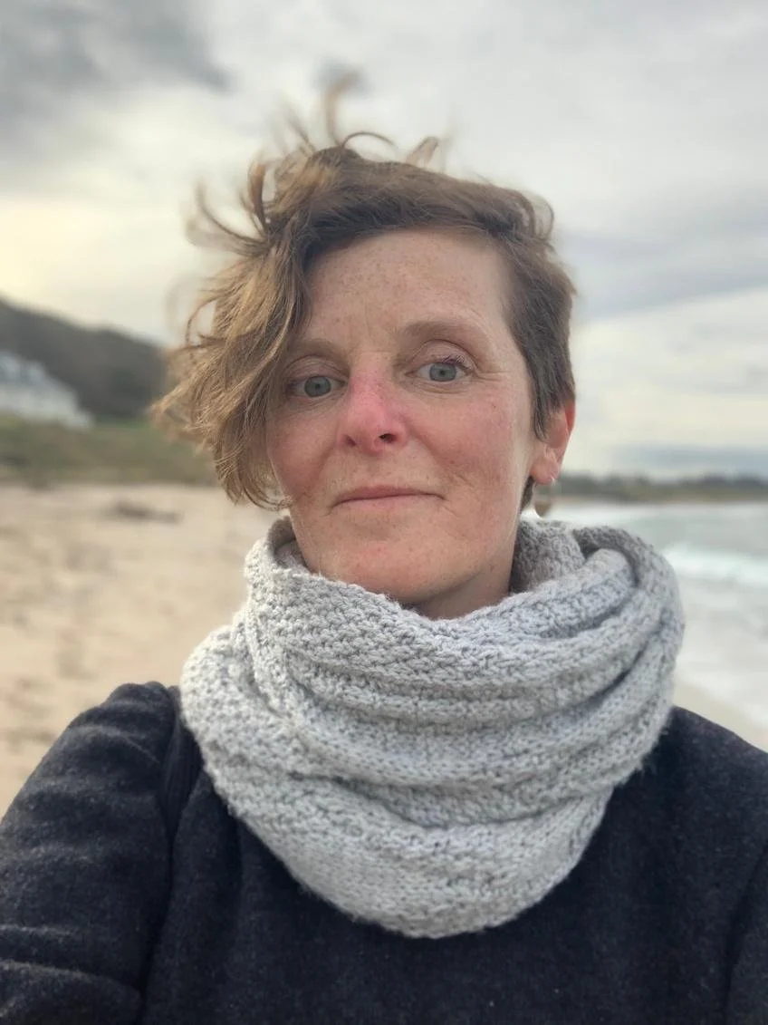 A woman with short, messy brown hair and pale skin taking a selfie on a beach with gray skies.