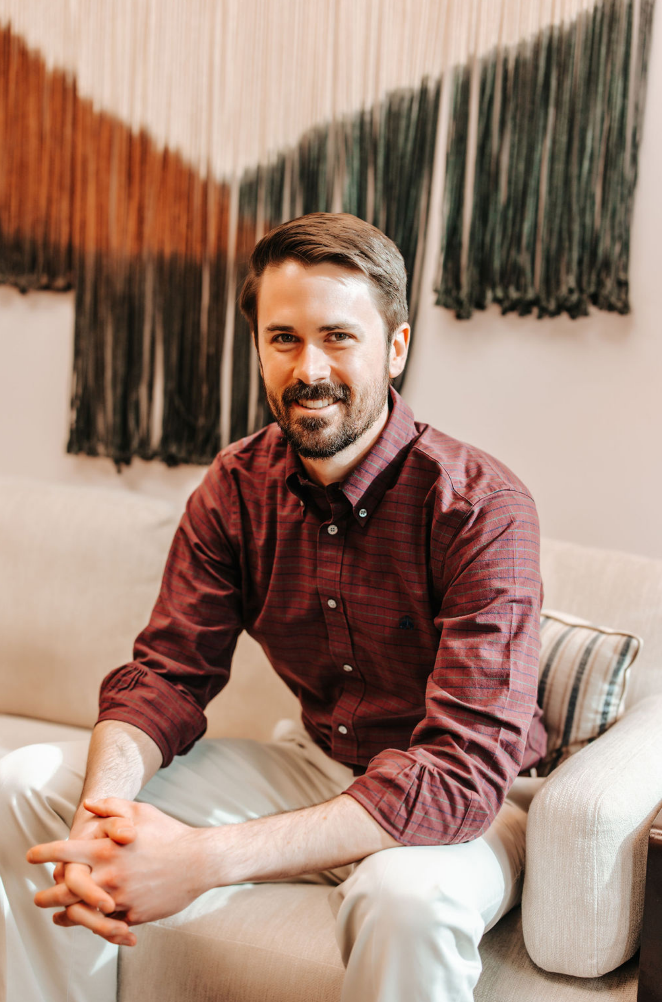 A man with a beard and mustache, wearing a maroon checkered shirt and beige pants, sitting on a beige sofa in front of woven textile wall art.