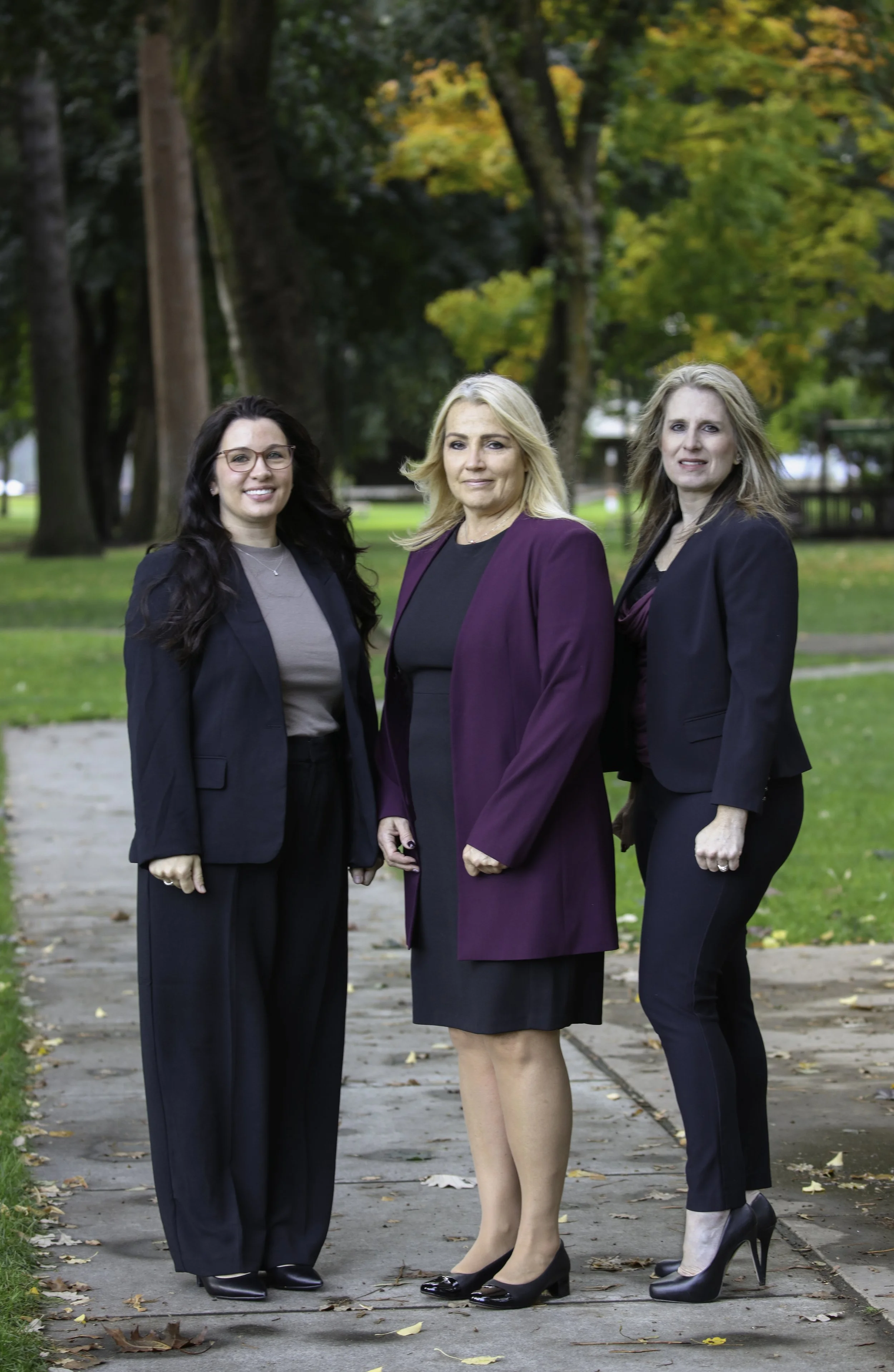 Three women standing on a park sidewalk, dressed in business attire, with trees and fall foliage in the background.