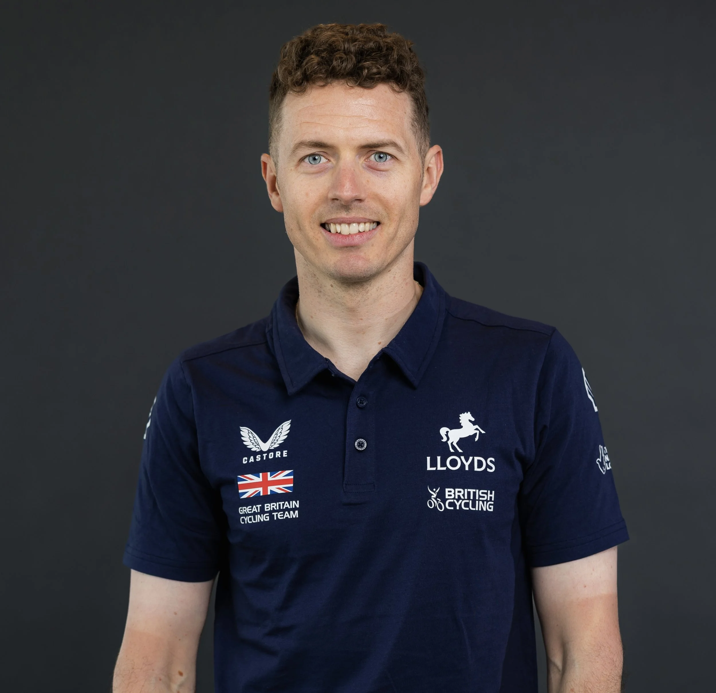 A man with short curly brown hair and blue eyes wearing a navy blue polo shirt with logos of British cycling teams and sponsors, standing against a dark gray background.