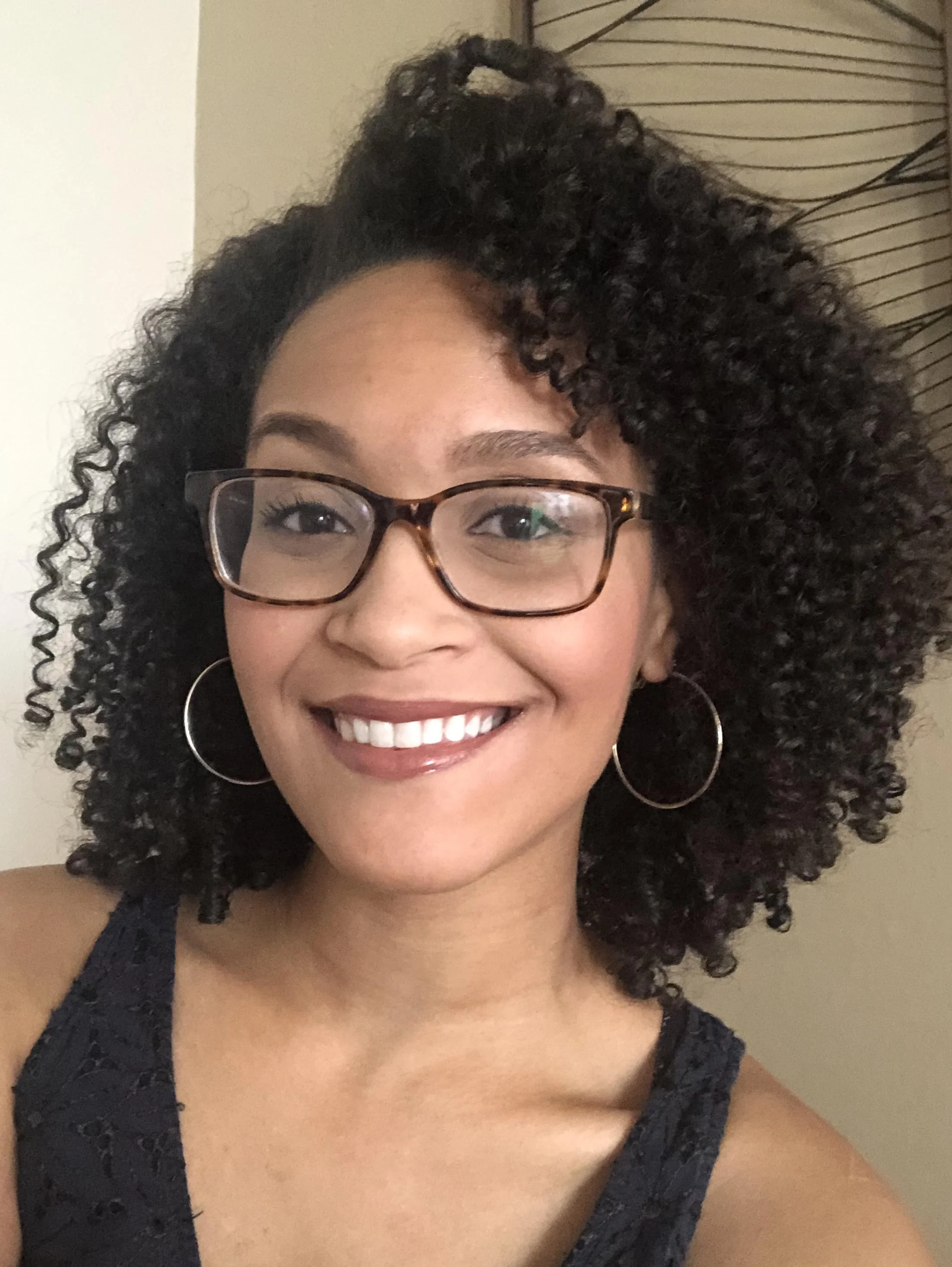 Smiling woman with curly hair, glasses, and hoop earrings taking a selfie indoors.
