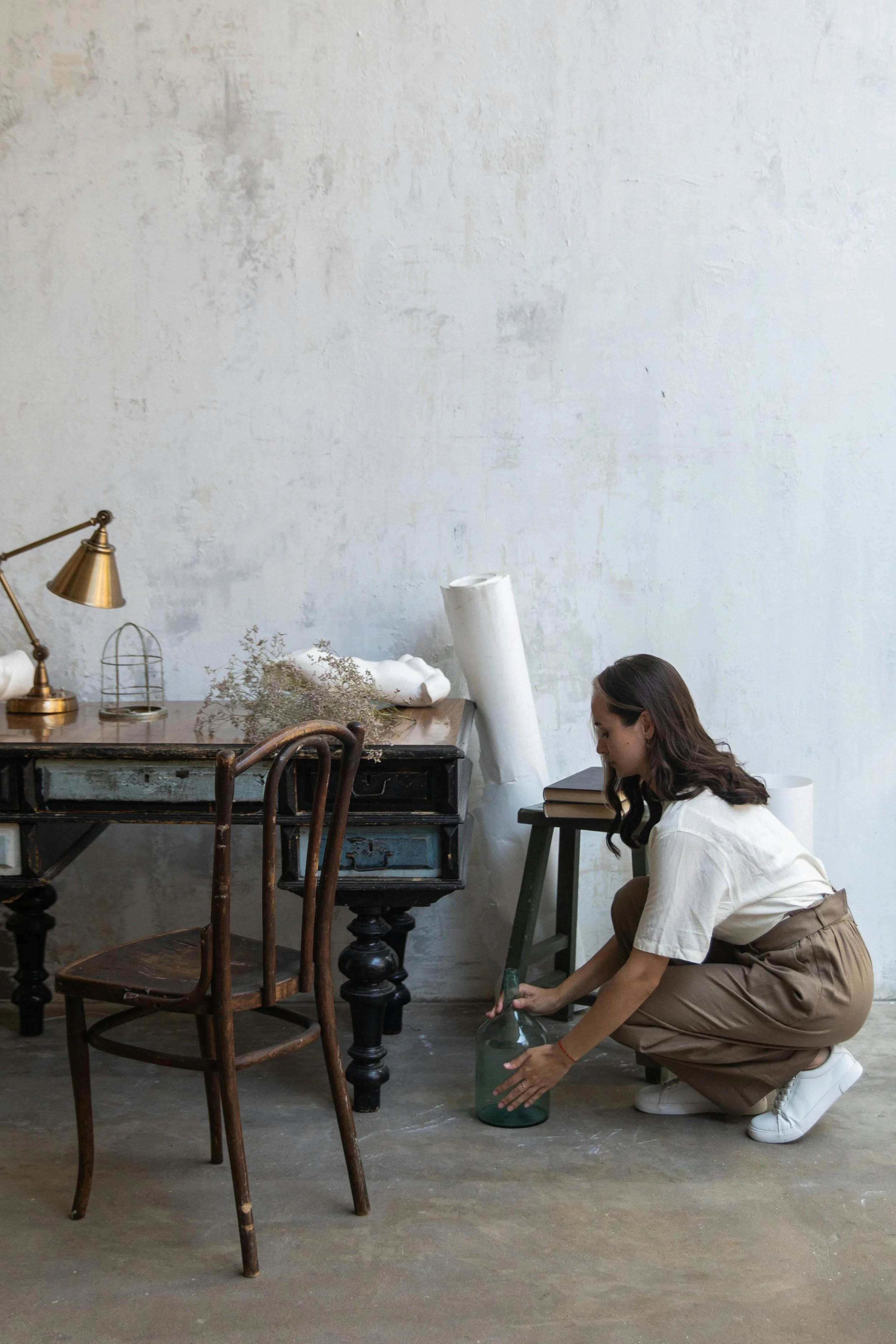 A woman crouching down placing a glass bottle on a concrete floor in a room with a vintage desk, chairs, and art supplies.