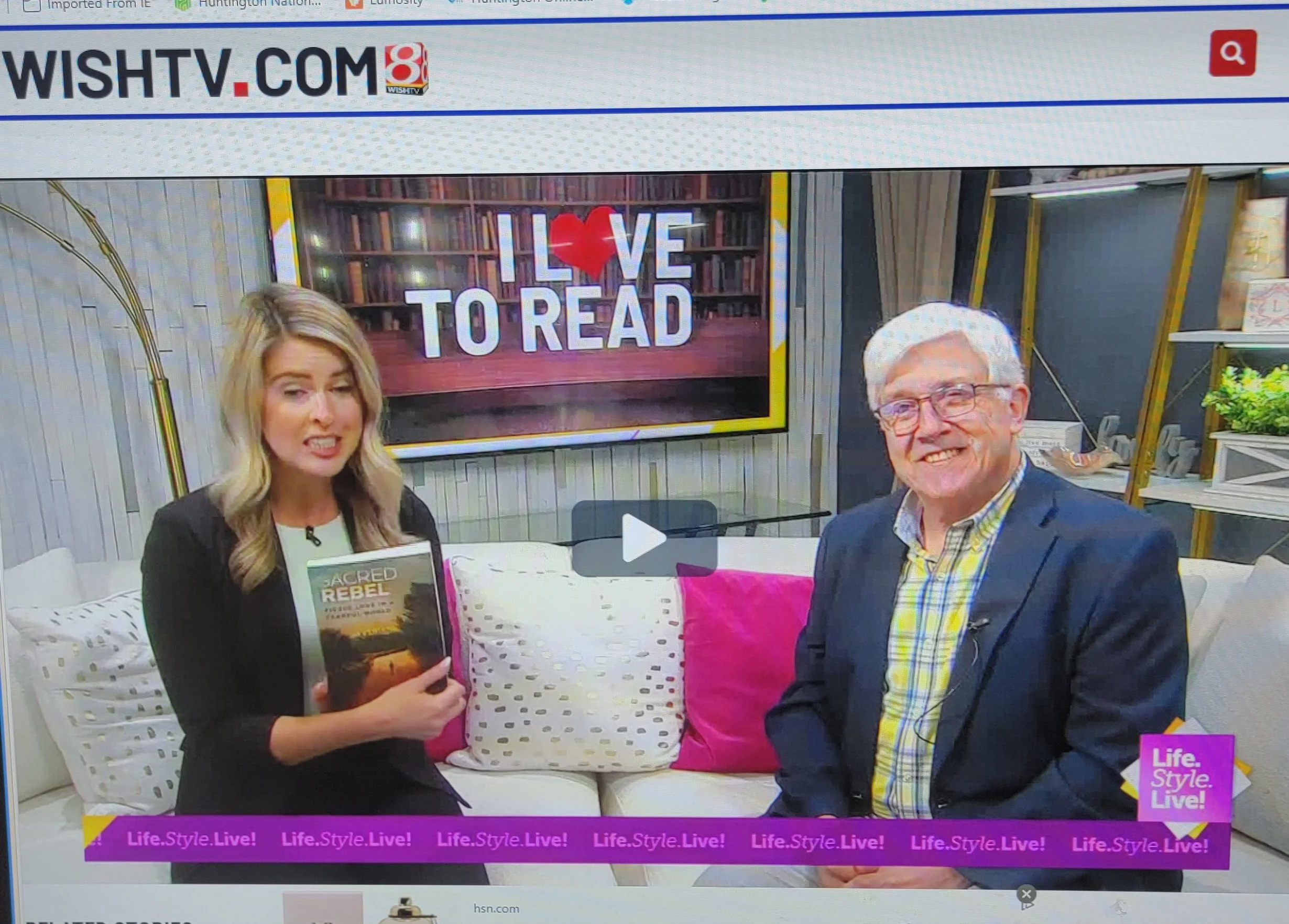 Two women sitting on a white couch in a room decorated with bookshelves, pillows, and plants, with a large TV screen behind them displaying the text 'LOVE TO READ' with a red heart. The woman on the left is holding a book titled 'Sacred Rebel,' and the woman on the right is smiling, wearing glasses, and a blazer. The bottom of the image has a pink banner with the text 'Life.Style.Live!' repeated.