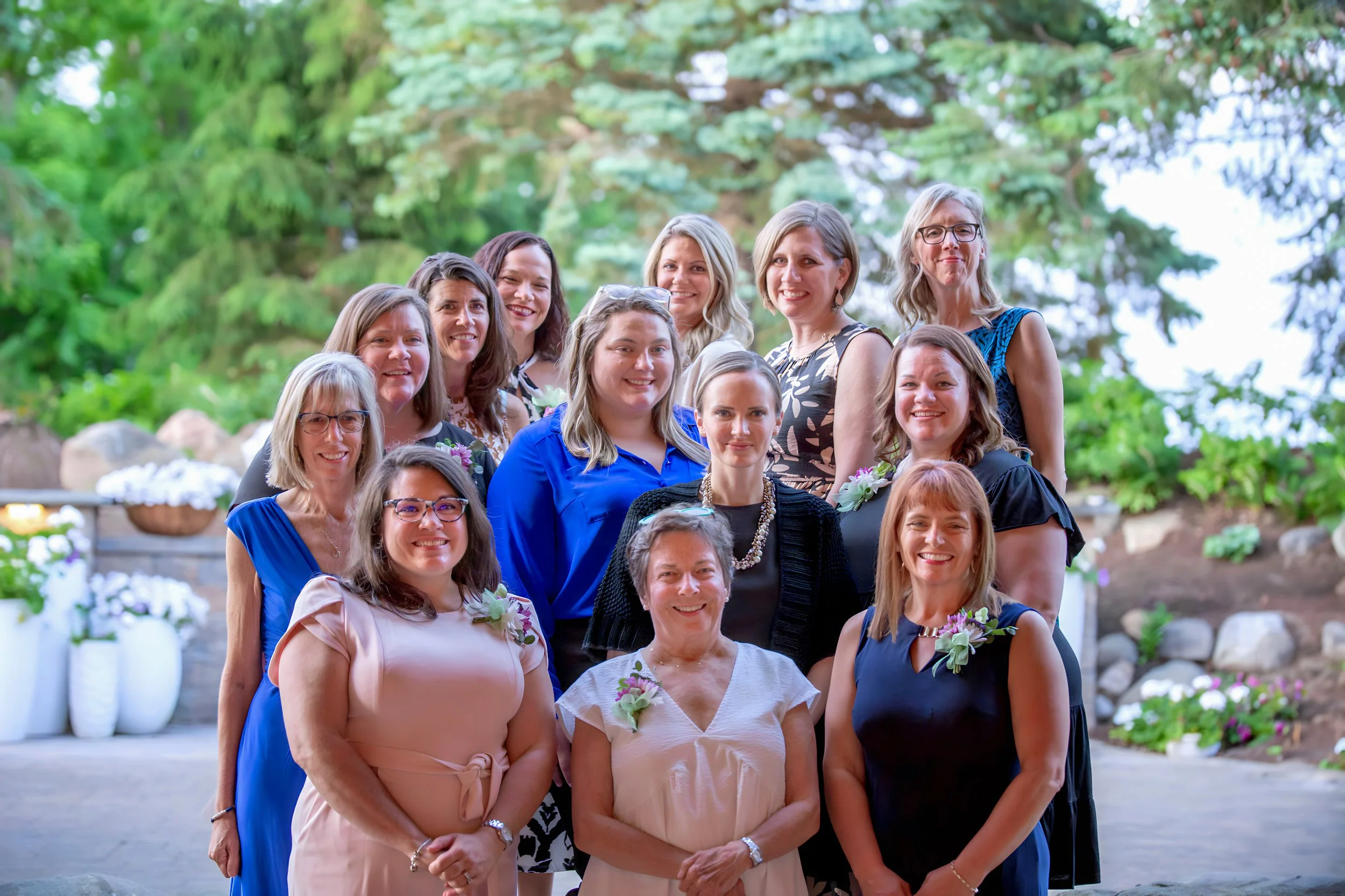 Group portrait of women dressed in formal attire, outdoors with greenery and flowers in the background, smiling at the camera.