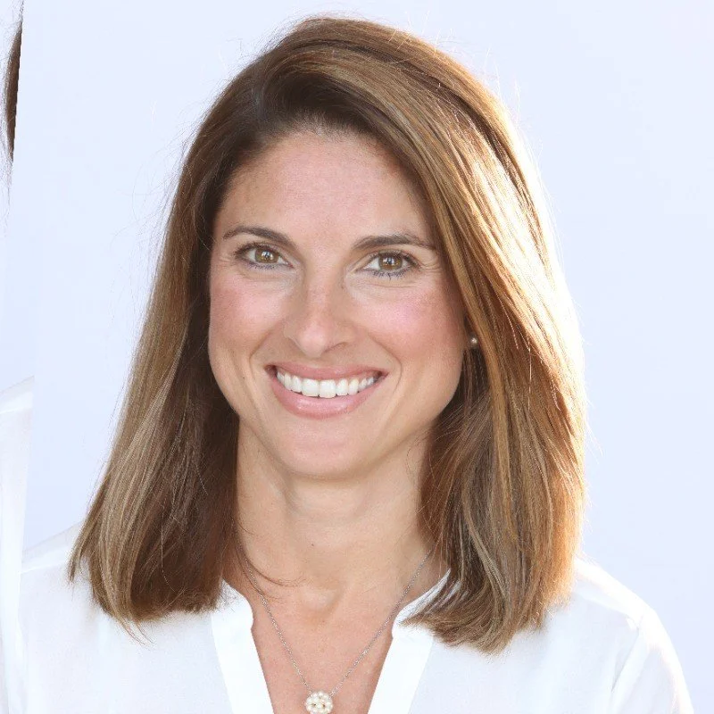 A smiling woman with shoulder-length brown hair and light skin, wearing a white top and a necklace.