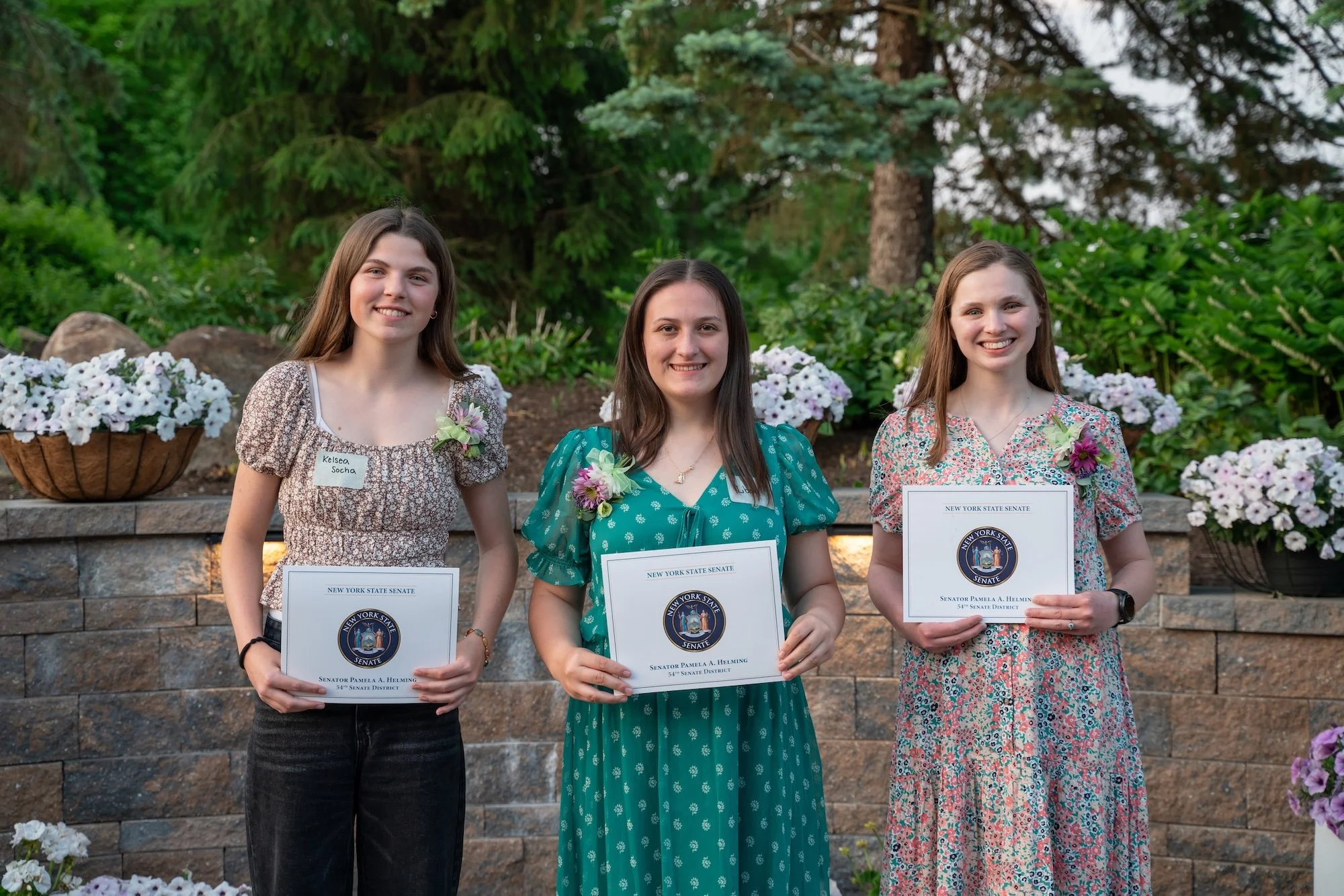 Three young women standing outdoors in front of a stone wall and garden, each holding a certificate, with floral decorations around them.