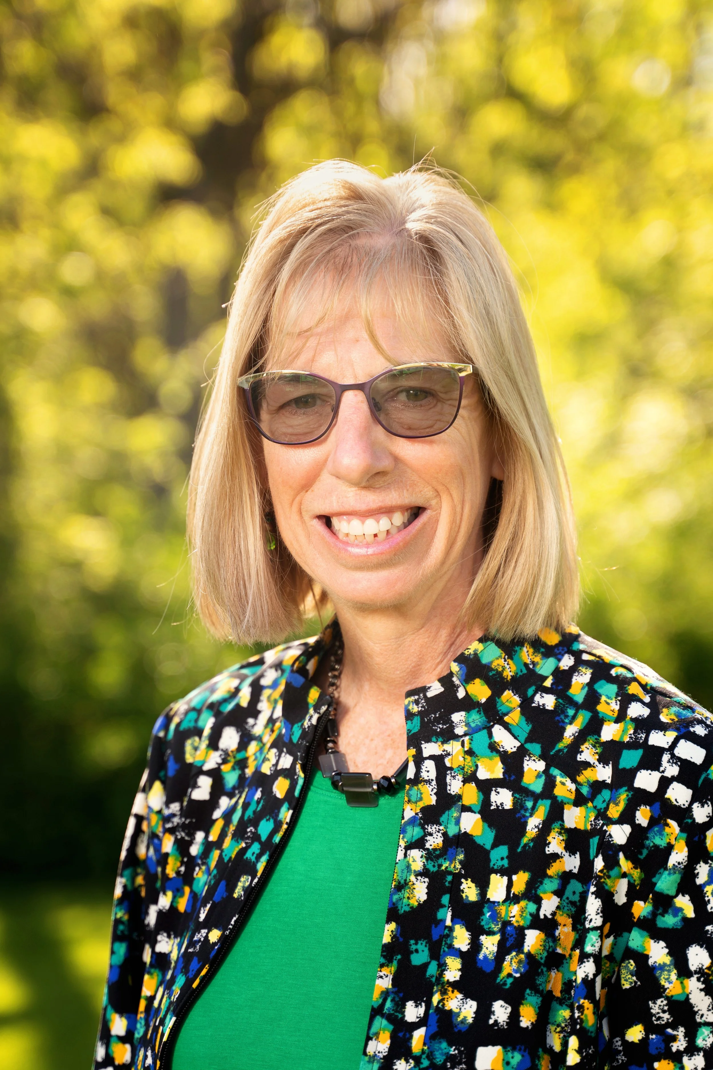 A smiling woman with blonde hair, wearing sunglasses, a green shirt, and a colorful patterned jacket, outdoors in a sunny, green park.