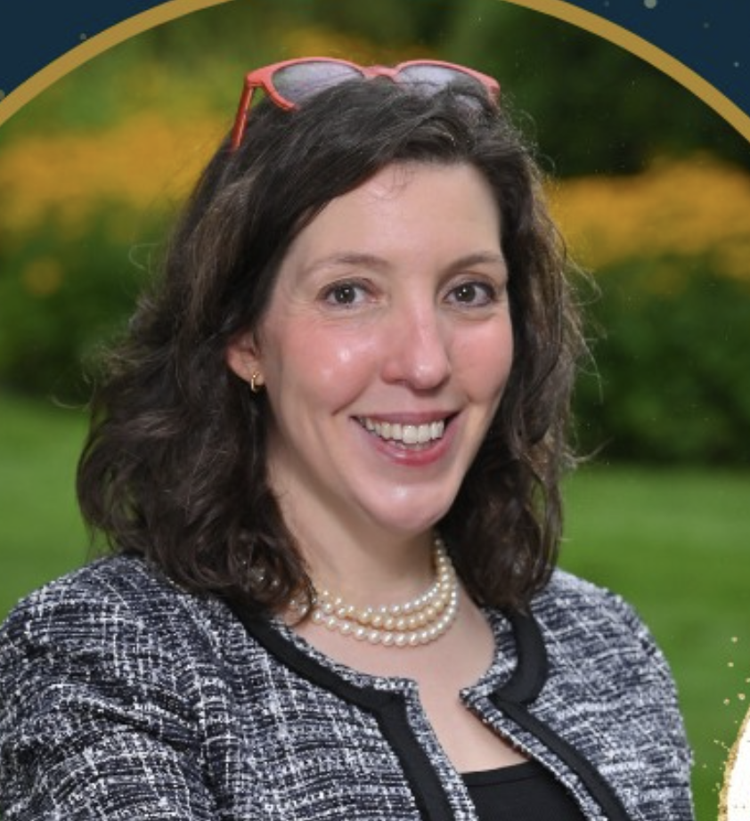 A woman with dark, curly hair, wearing pink glasses on her head, pearl necklace, earrings, and a patterned blazer, smiling outdoors with a colorful blurred background.