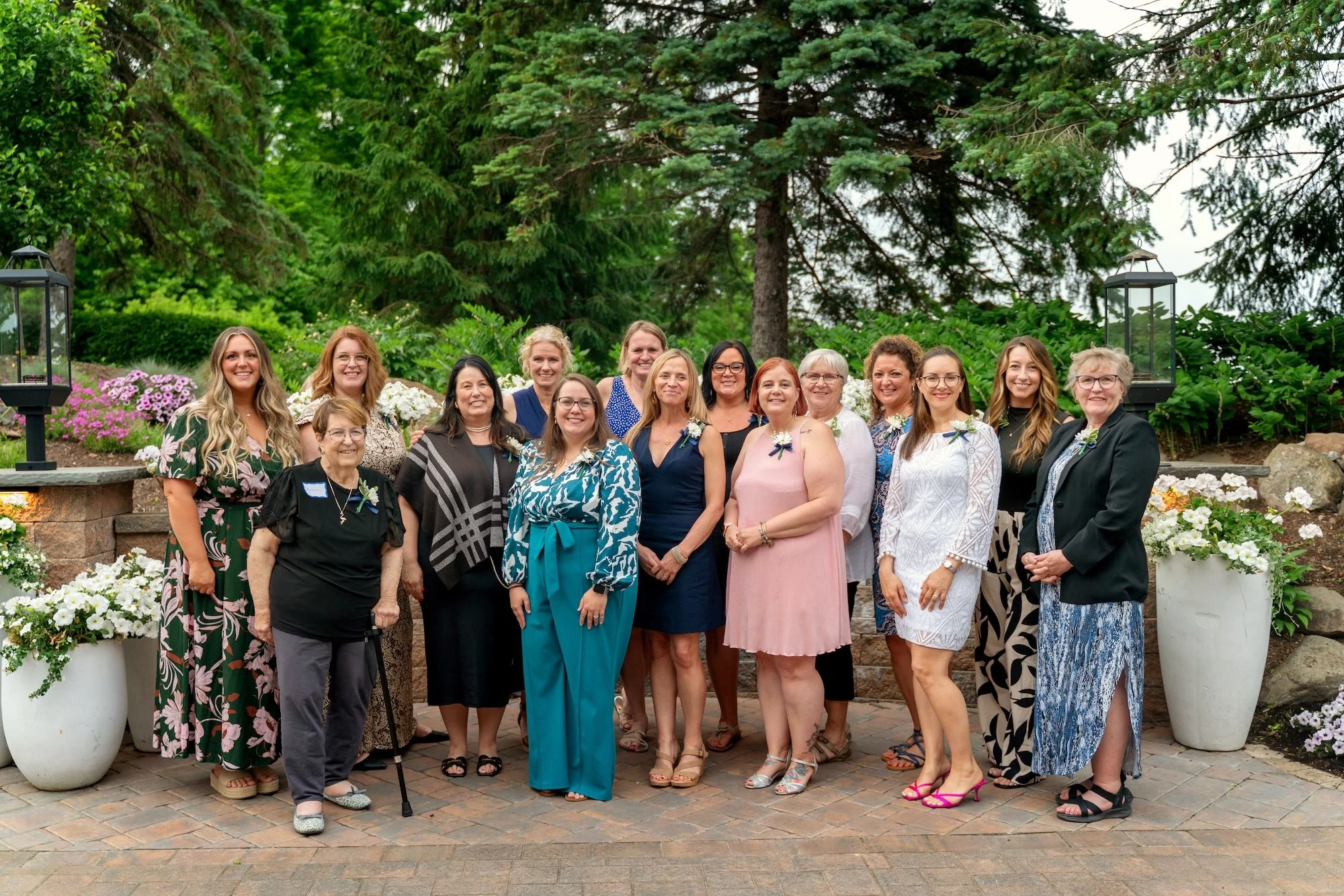 Group of women standing outdoors in a garden with trees and flowers, posing for a photo on a brick pathway.