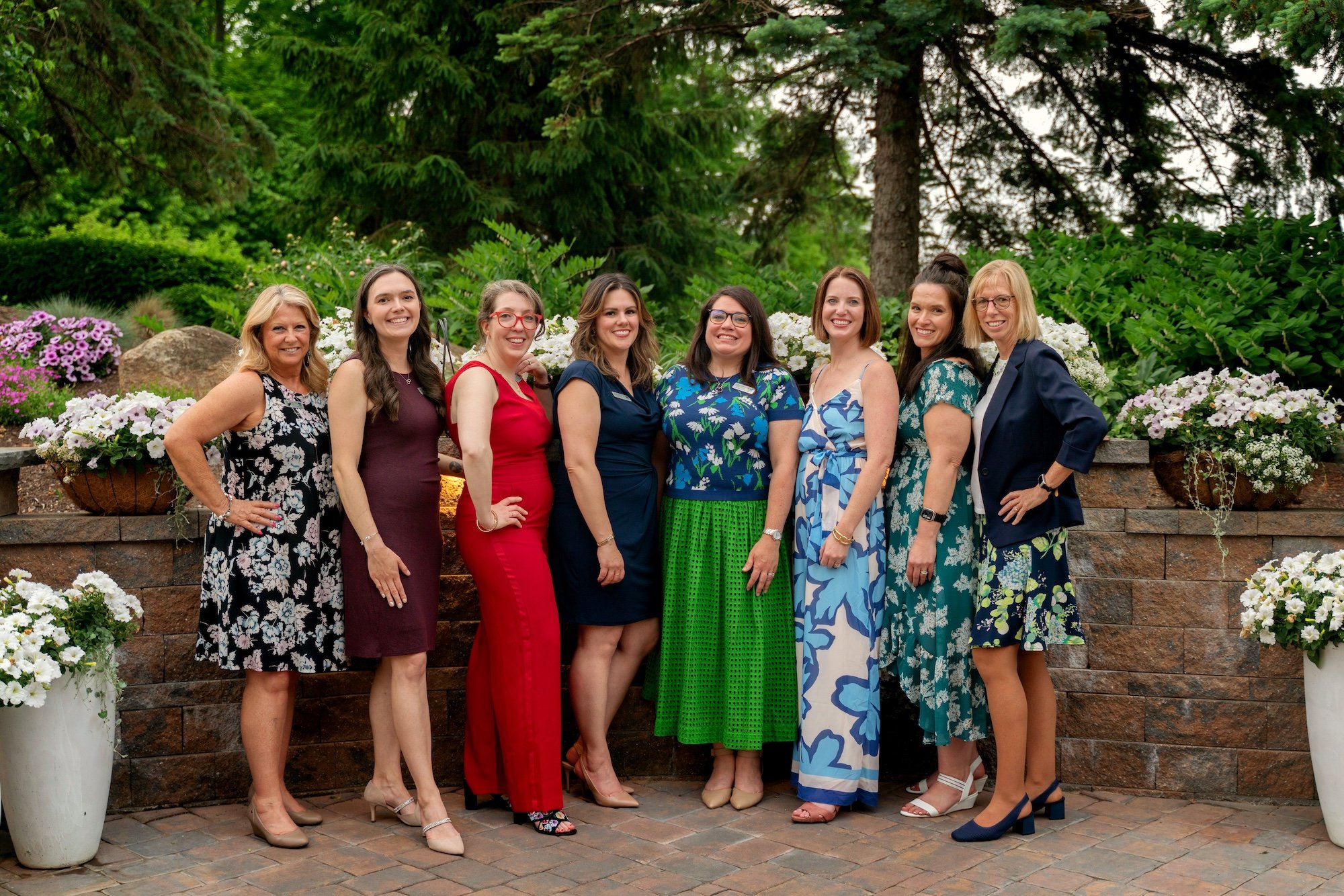 Group of women standing together outdoors in front of a garden with flowers and trees, posing for a photo.