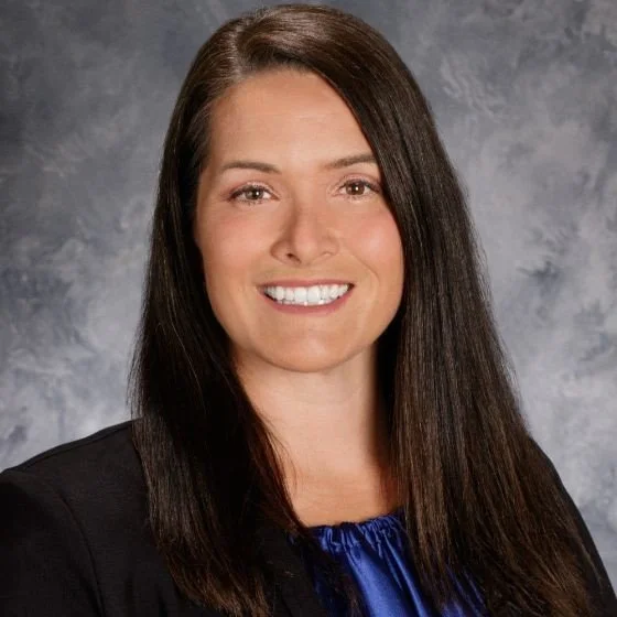 A professional woman with long dark hair, wearing a black blazer and a blue blouse, smiling in front of a gray mottled background.