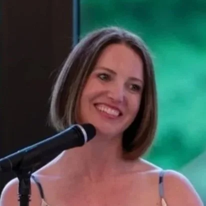 A woman with shoulder-length brown hair smiling in front of a green background, speaking into a microphone.