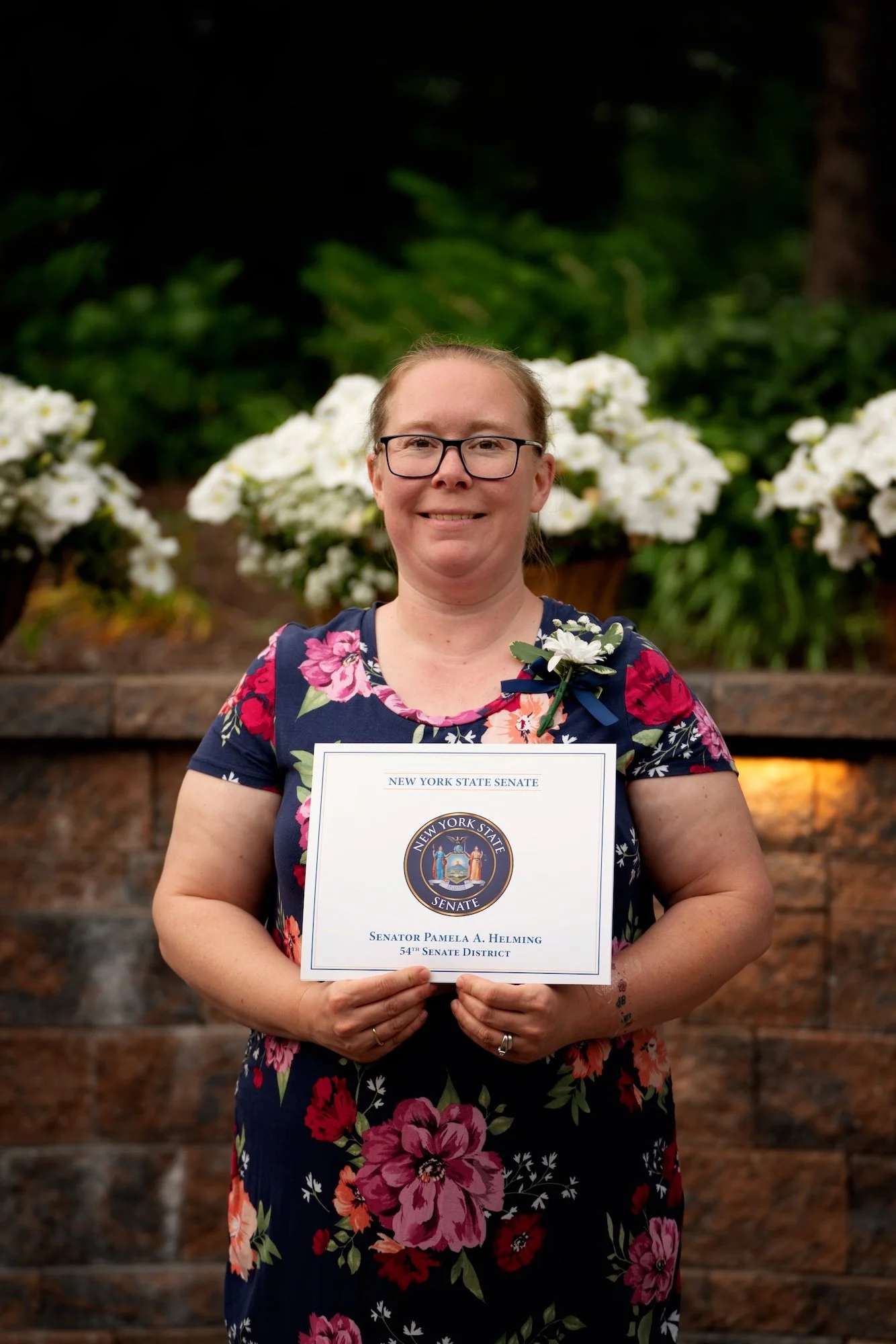 Woman in floral dress holding certificate, standing outdoors with white flowers and greenery in background.