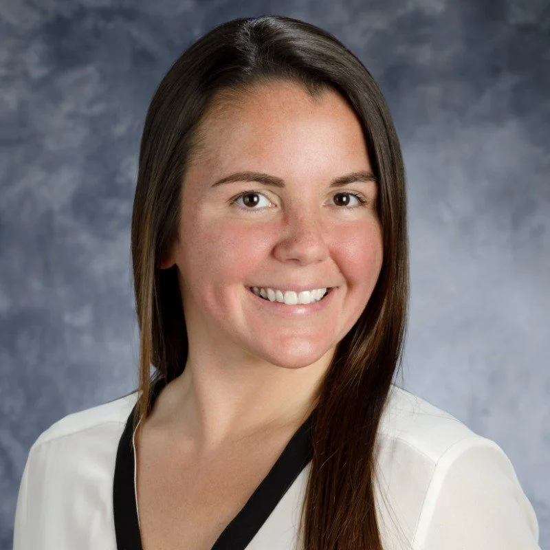 Professional portrait of a woman with long brown hair, wearing a white top with black trim, smiling against a gray mottled background.
