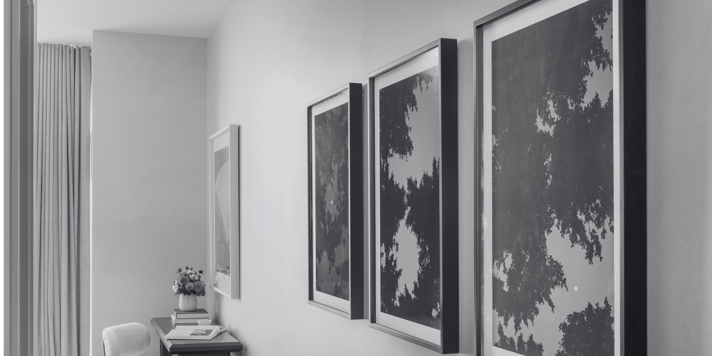 Black and white photograph of three framed pictures hanging on a white wall in a room with a desk, a chair, books, a vase with flowers, and a window with white curtains.