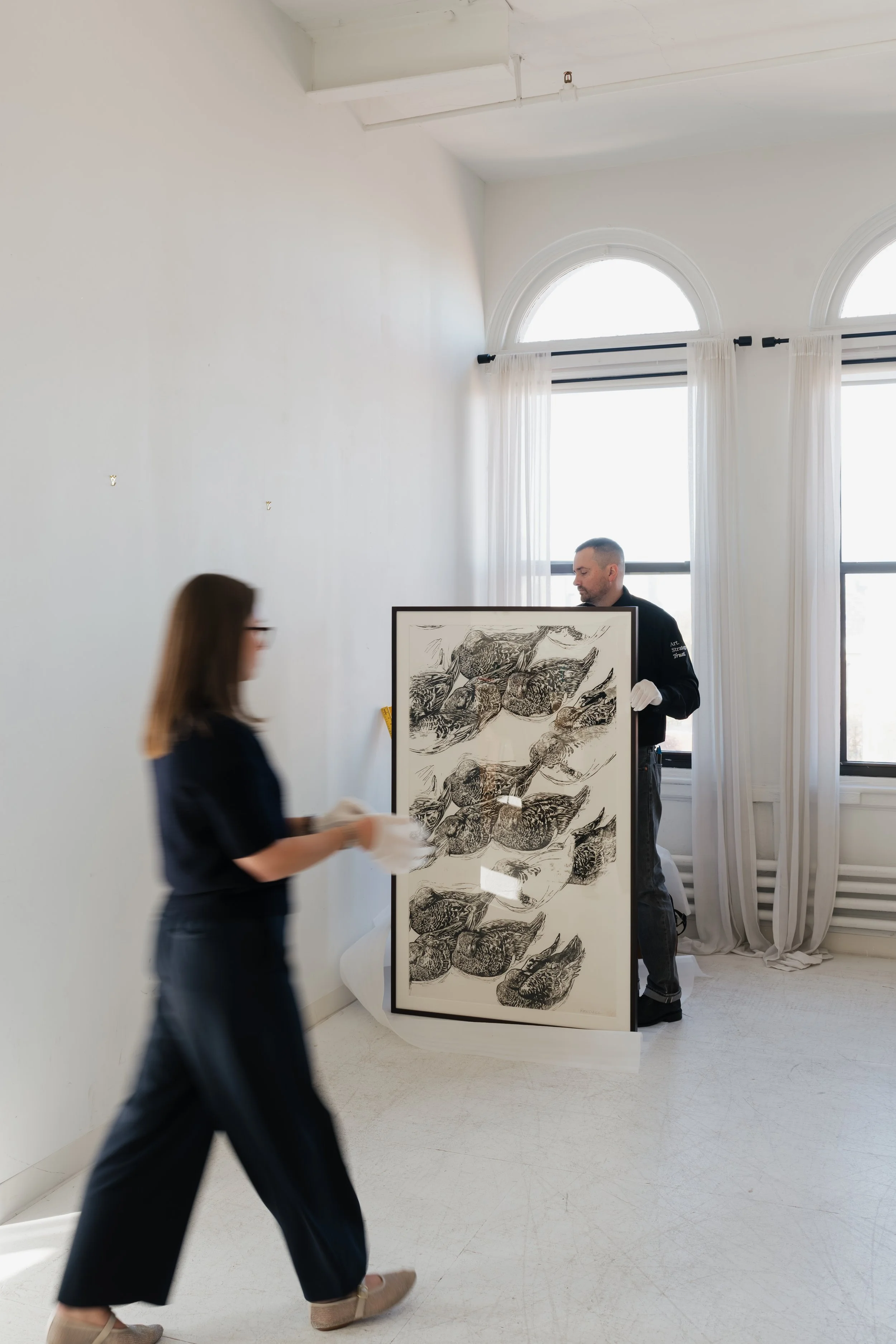 Two people handling a framed artwork or print in a bright art gallery or museum with large arched windows and white curtains.