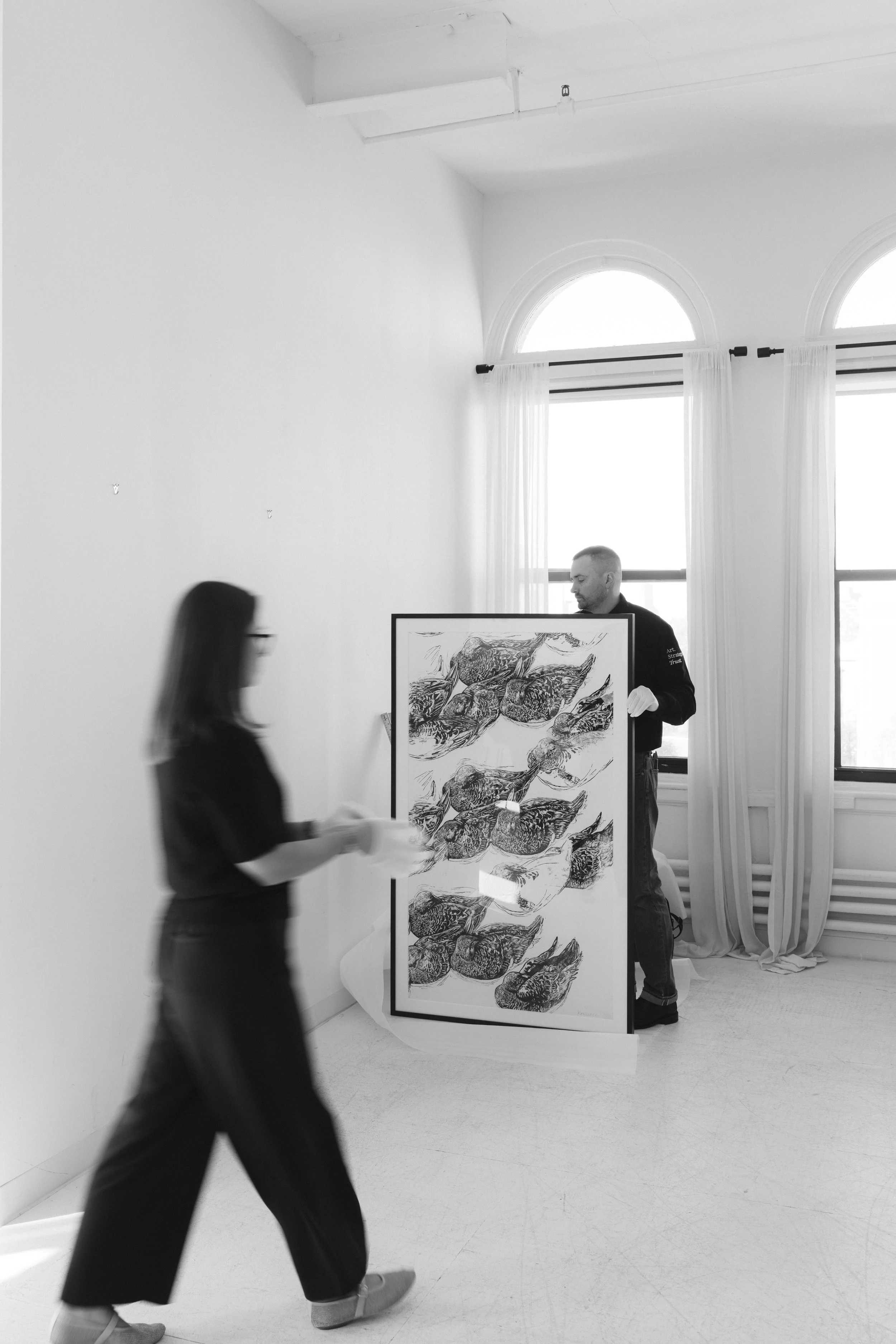 Two people in a gallery setting, one man holding a framed artwork of multiple bird sketches, with a woman walking past in front of him, all in black and white.