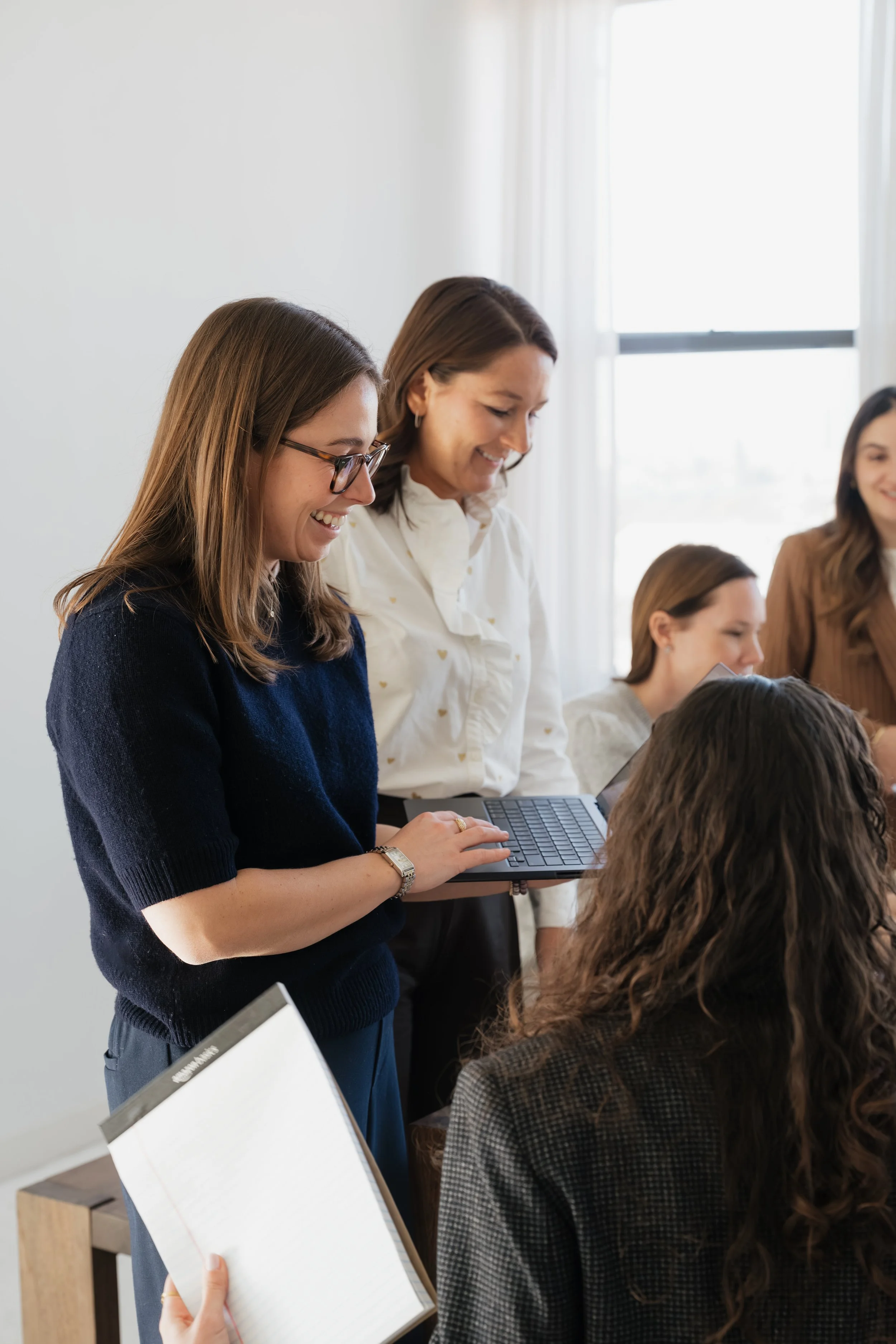 A group of women standing and sitting in a meeting room, smiling and talking to each other, with one woman holding a laptop and another holding a notebook.