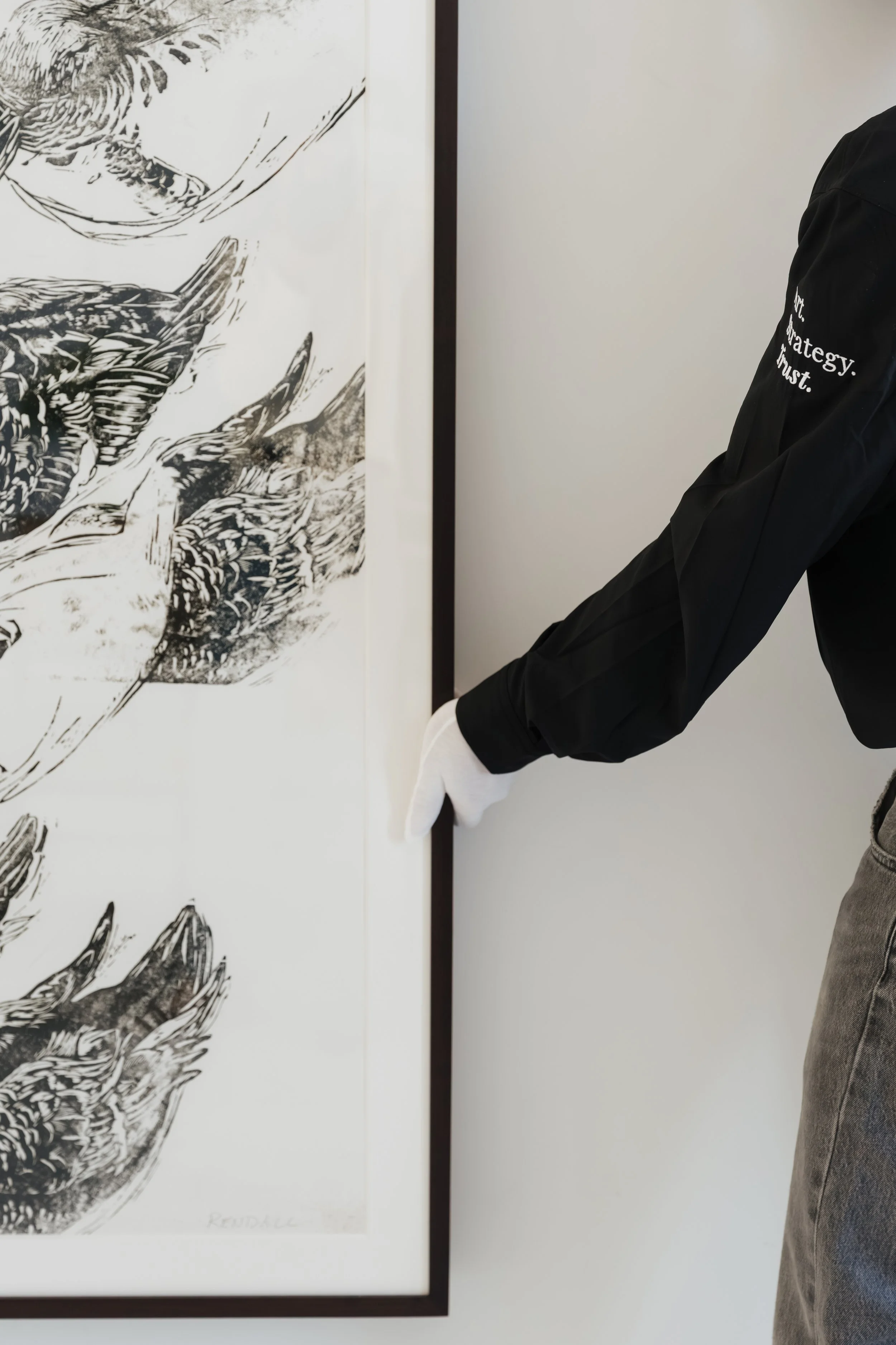 Person wearing white glove adjusting a framed black and white artwork on a white wall.