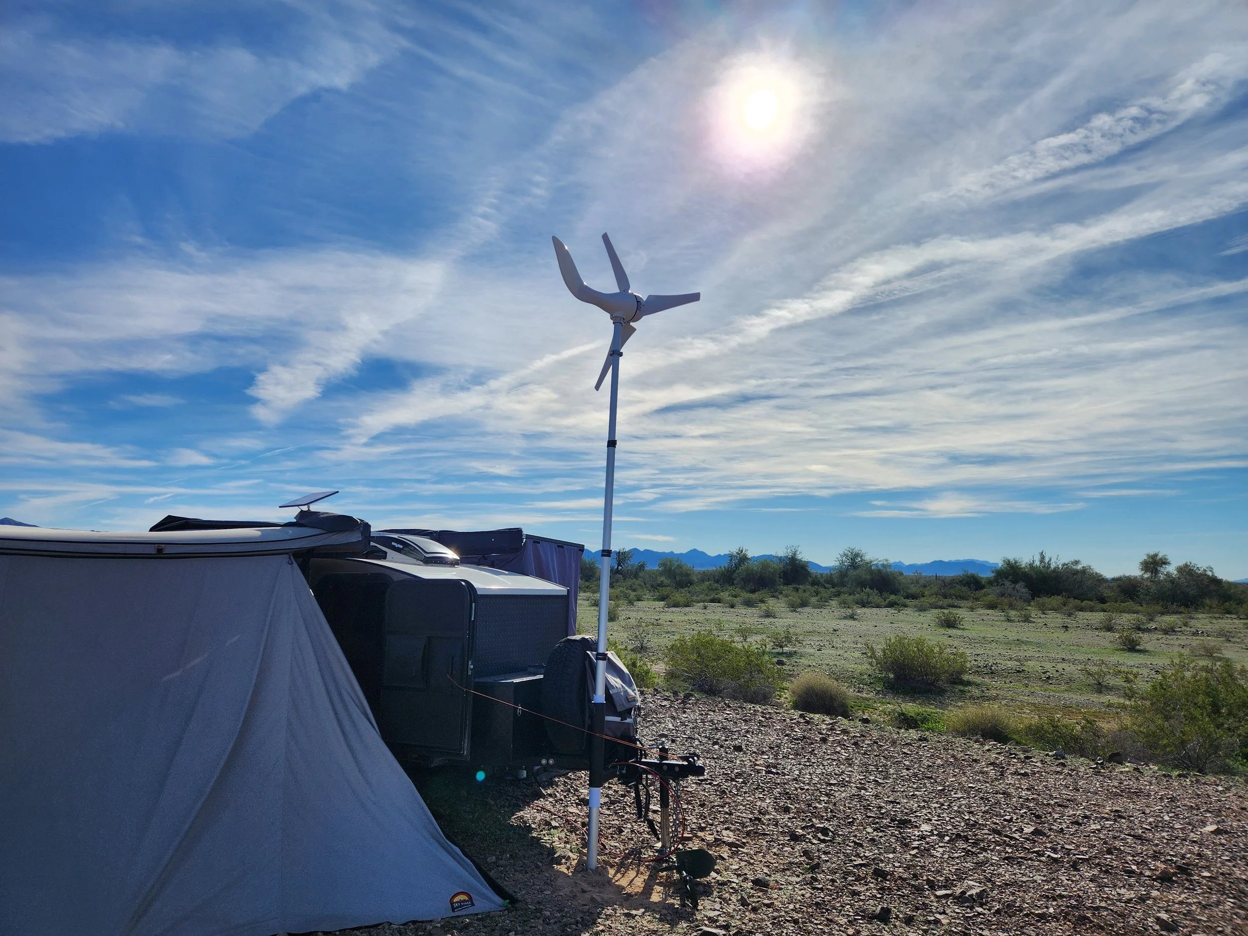 A camping scene in a desert with a tent, a wind turbine, and a trailer, under a partly cloudy sky with the sun shining.