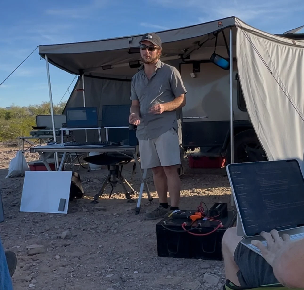 A man standing outdoors next to a camper trailer, wearing sunglasses, a hat, and casual clothing, surrounded by equipment and screens.