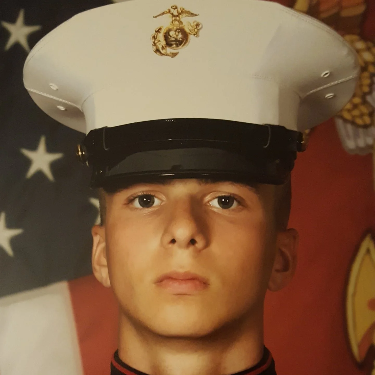A young boy in a U.S. Marine dress uniform with a white dress cap featuring the Marine Corps emblem, against a background of American flags and military insignia.