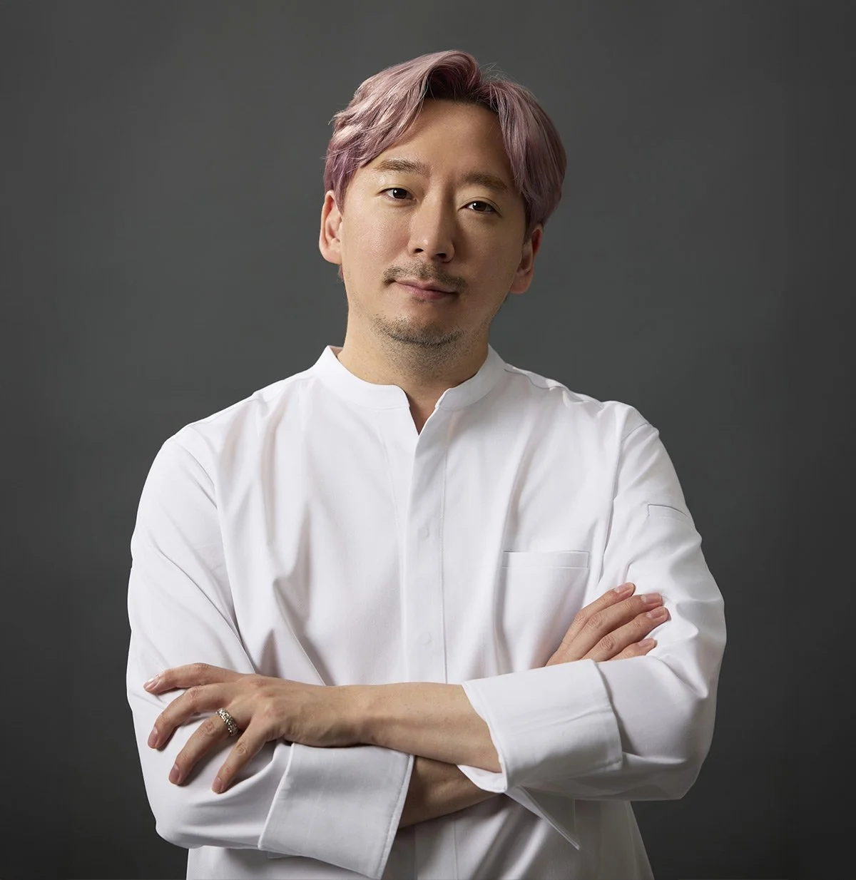 A smiling man with dark hair and a beard, wearing a dark gray shirt, standing indoors with a blurred background.