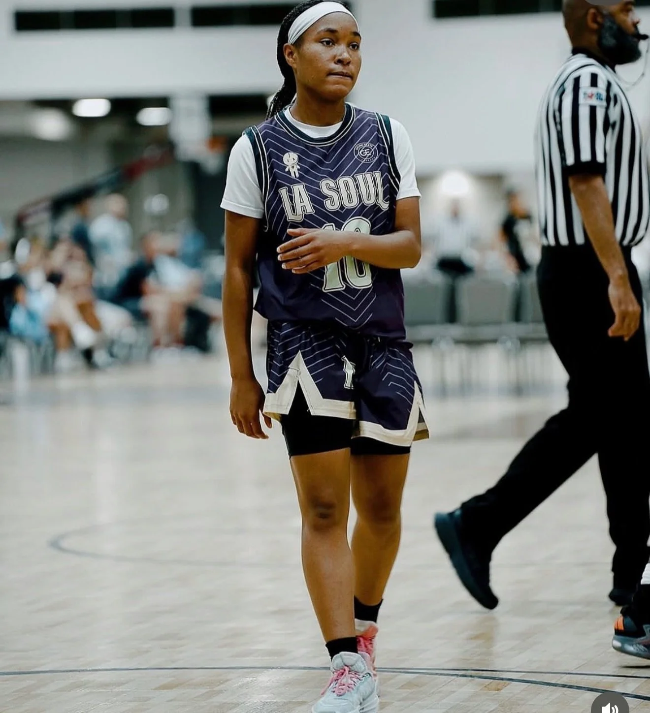 A female basketball player in a uniform with 'LA SOUL' written on it, standing on an indoor basketball court during a game, with spectators and a referee in the background.