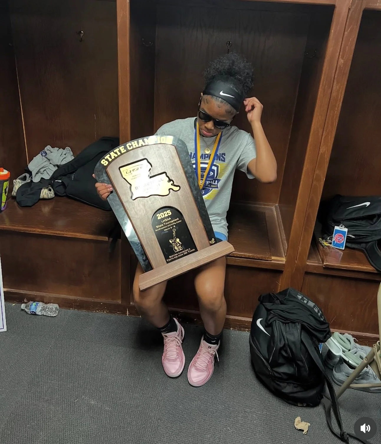 A young female athlete sitting in a locker room holding a large sports trophy shaped like the state of Louisiana. She is wearing a gray Champion T-shirt, pink sneakers, sunglasses, and a headband, with a medal around her neck. The locker room has wooden lockers and various personal items scattered around.
