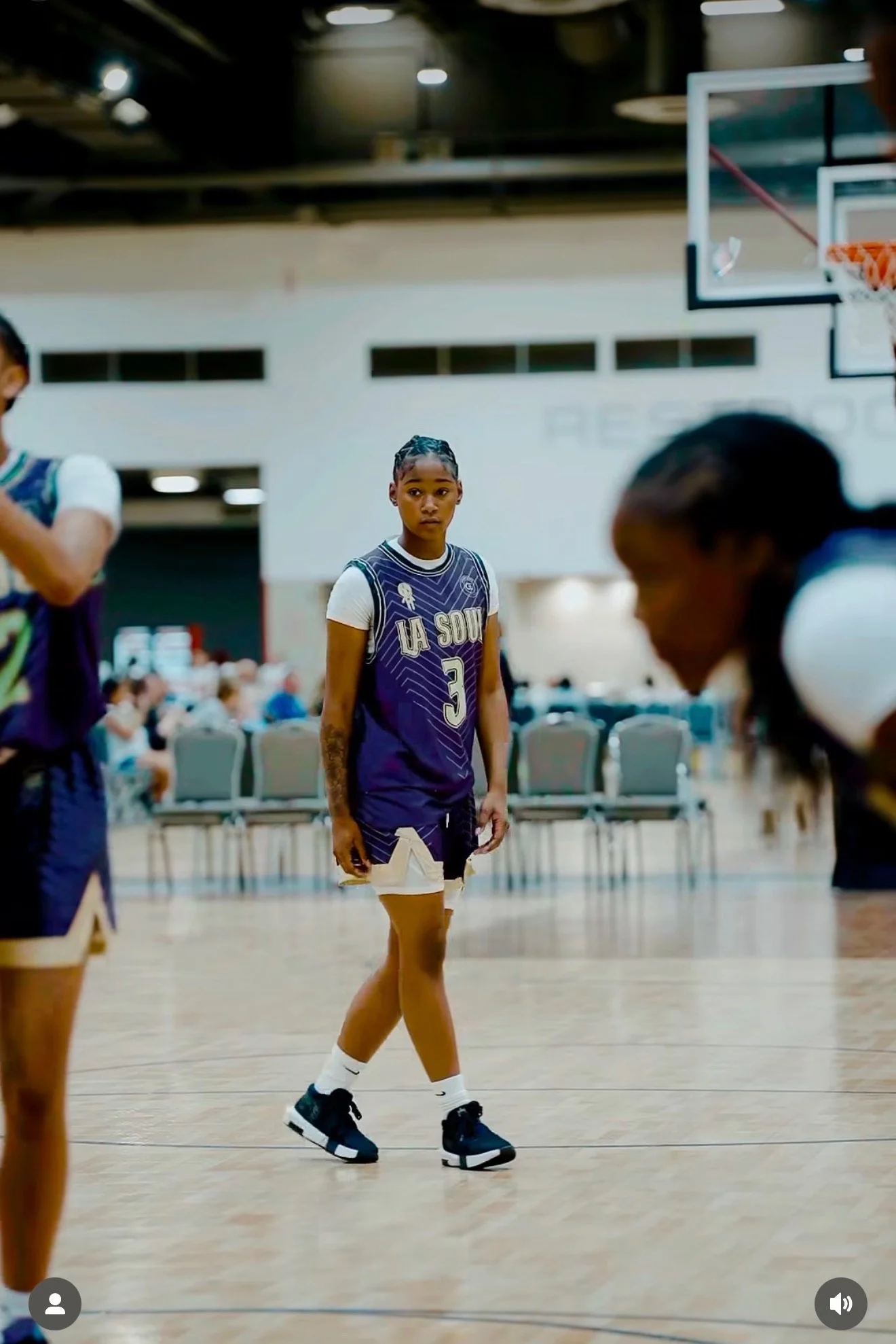 A young female basketball player standing on a basketball court during a game or practice, wearing a purple jersey with "LA SOU" and the number 3, with an attentive expression.