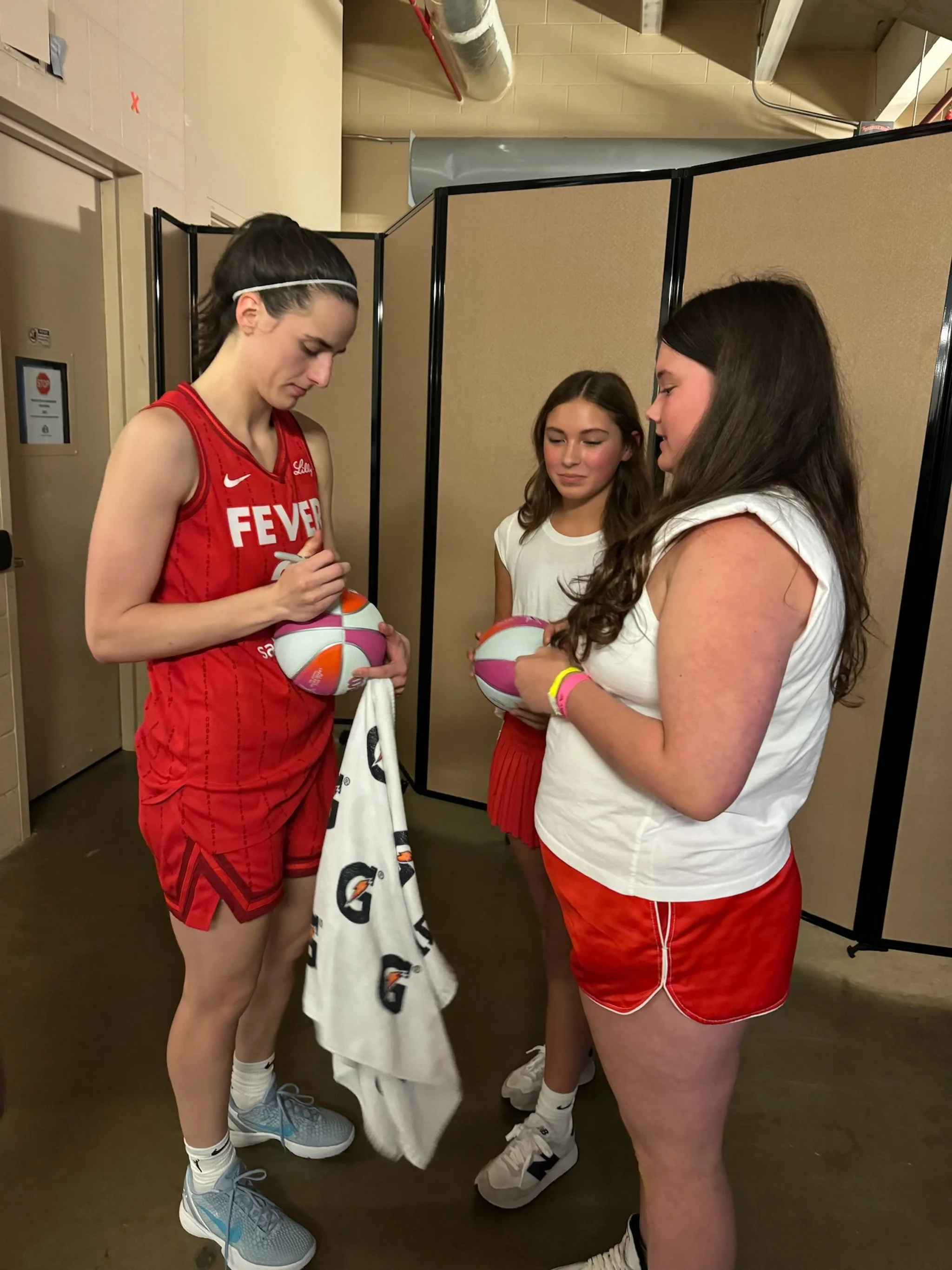 Three young female basketball players in red and white uniforms signing autographs and holding basketballs backstage.