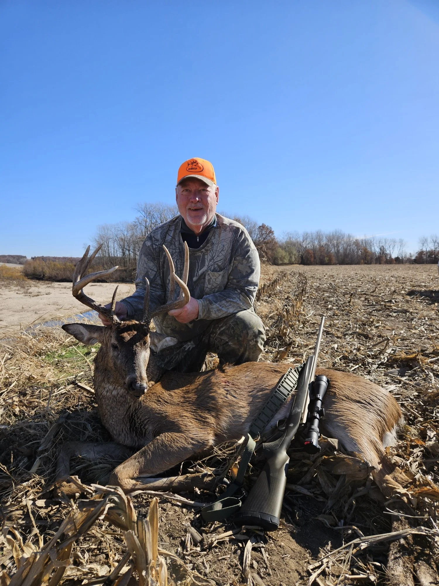 A man dressed in camouflage clothing kneels on a harvested field next to a large deer with antlers. He is holding the deer's antlers and smiling at the camera. A rifle with a scope is resting on the ground next to the deer against a backdrop of clear blue sky and leafless trees.