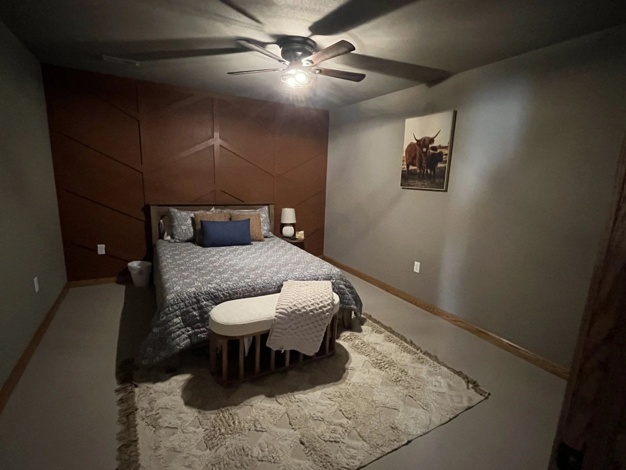 A cozy bedroom with a wooden headboard, a quilted bed, pillows, a small bench at the foot of the bed, a carpet rug, a wall painting of cattle, and a ceiling fan with lights.