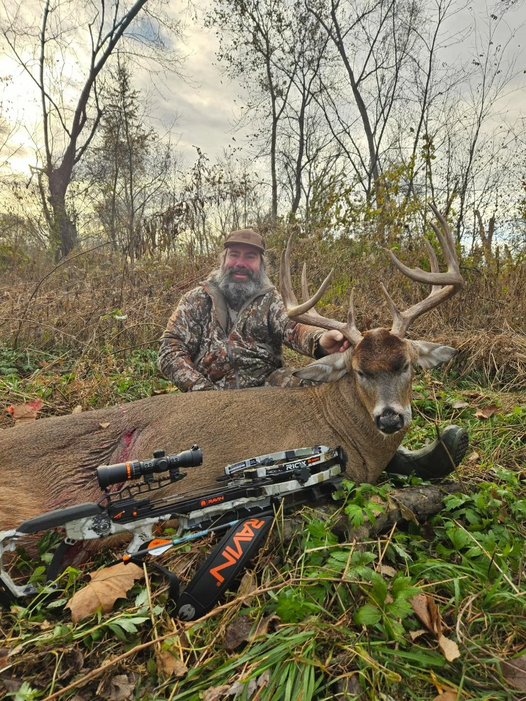 A man in camouflage clothing with a beard and a cap sitting next to a large deer with antlers, on a forest floor with a hunting rifle and scope placed on the ground nearby.