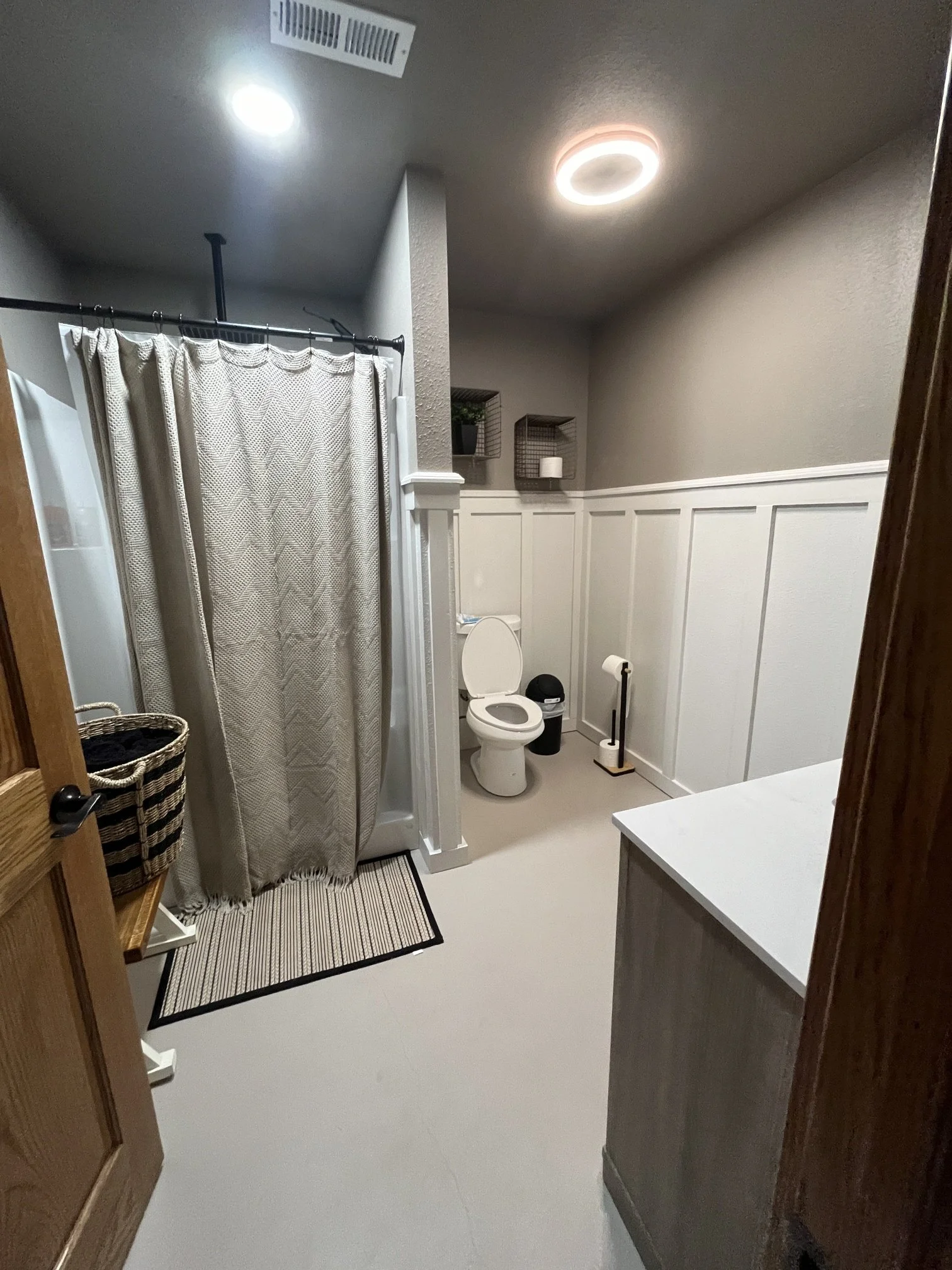 A bathroom with a shower and curtain, a toilet, a trash can, a toilet paper holder, shelves with a plant and a candle, and a ceiling light.