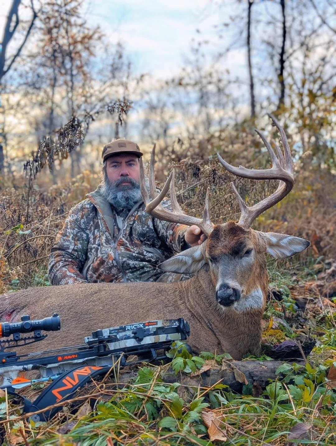 A man with a gray beard and camo clothing sitting in a forested area holding the antlers of a large buck with a bow resting in front of it.