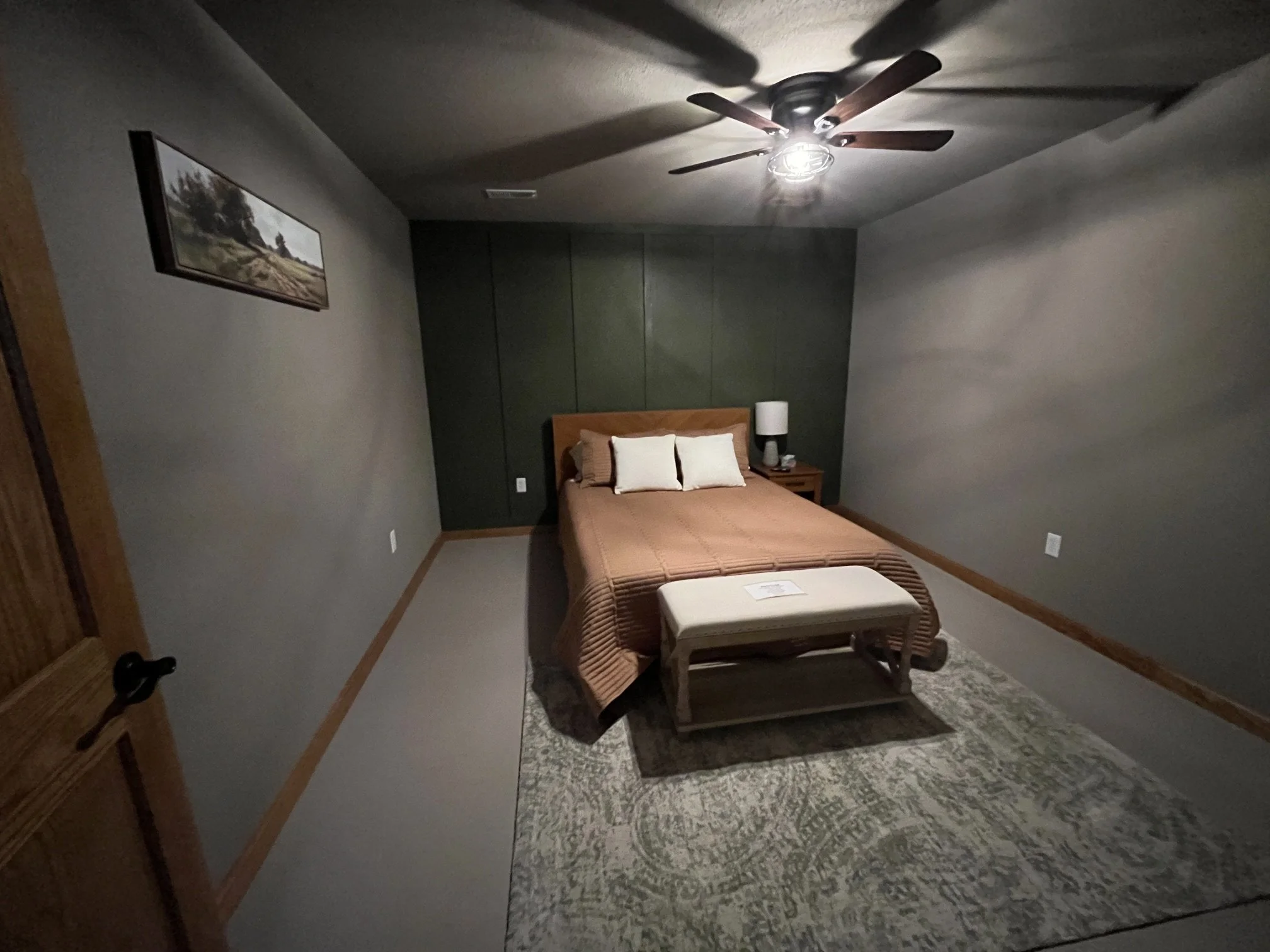 A bedroom with a bed, bench, side table with lamp, green accent wall, and wall art, lit by a ceiling fan light.