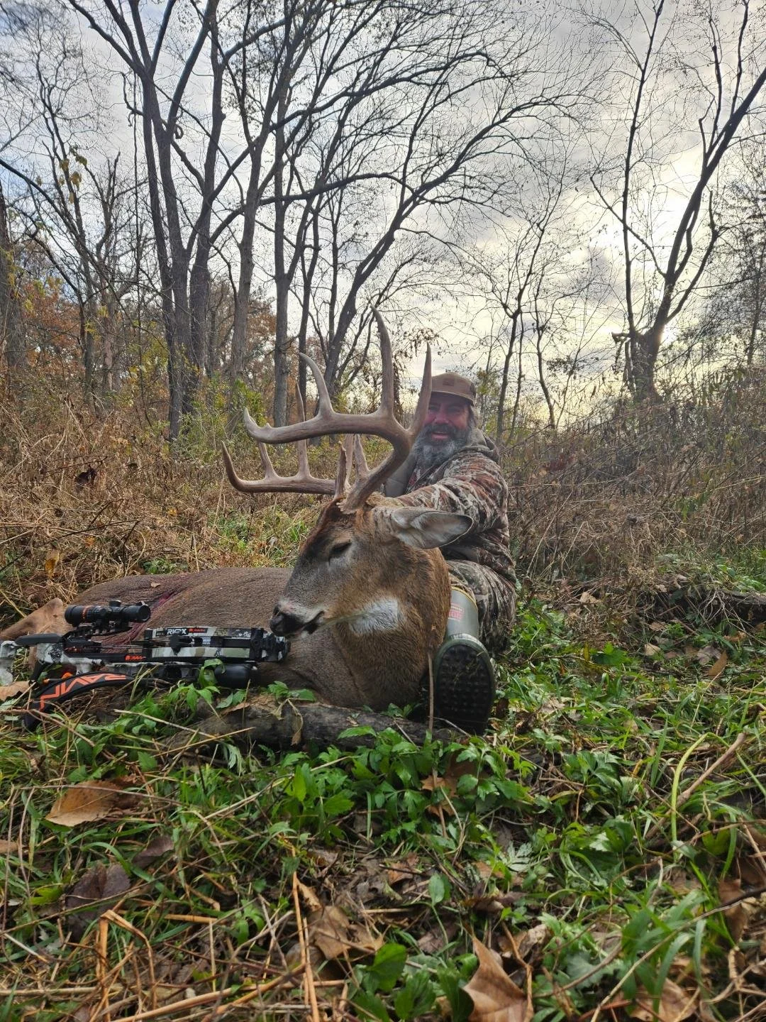A man in camouflage clothing sitting in a wooded area beside a large dead deer with antlers, with a rifle resting on the ground nearby.