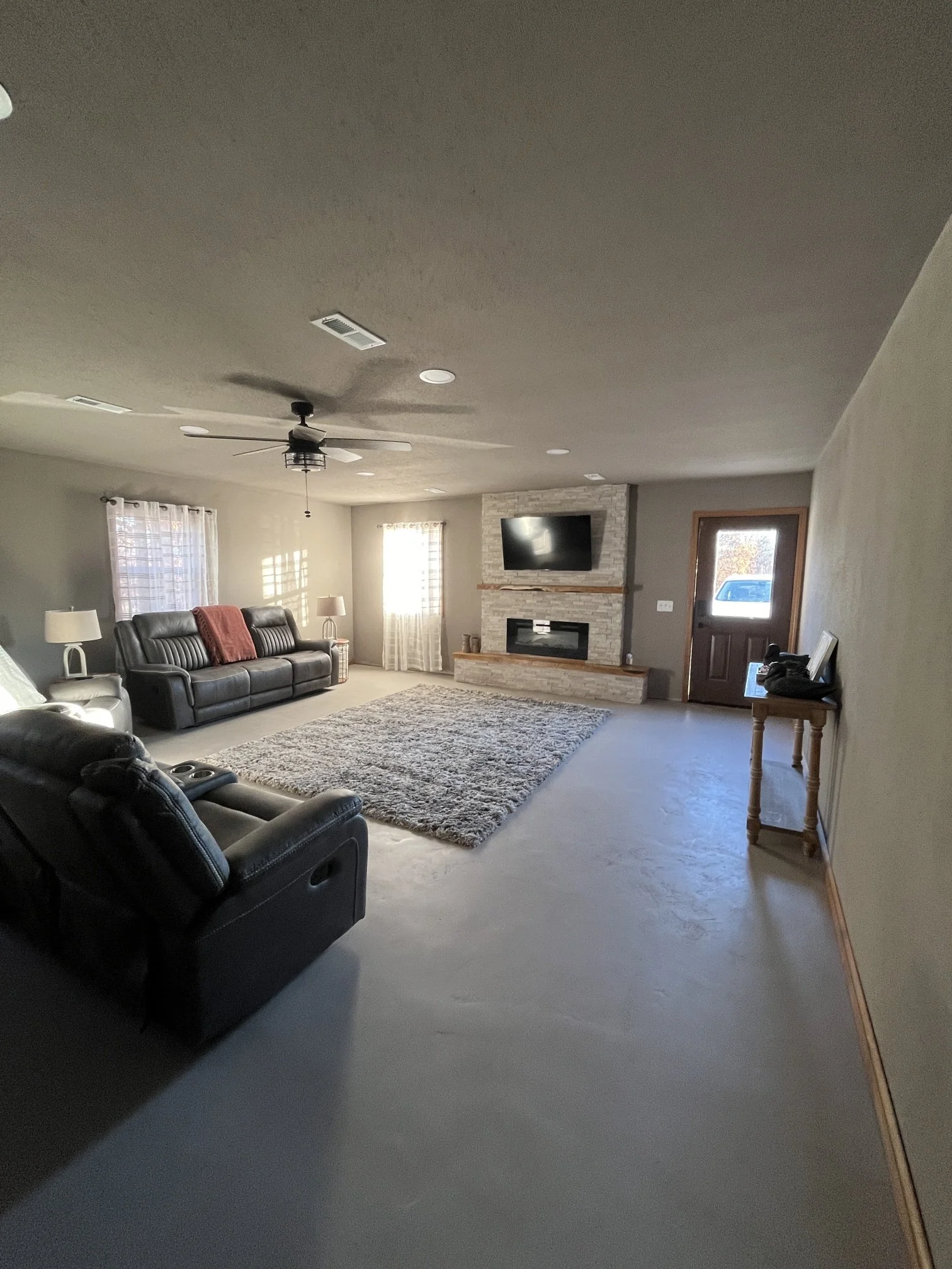 A living room with two black leather sofas, a rug, a fireplace with a mounted TV, beige carpet, and a door leading outside, with windows allowing natural light.