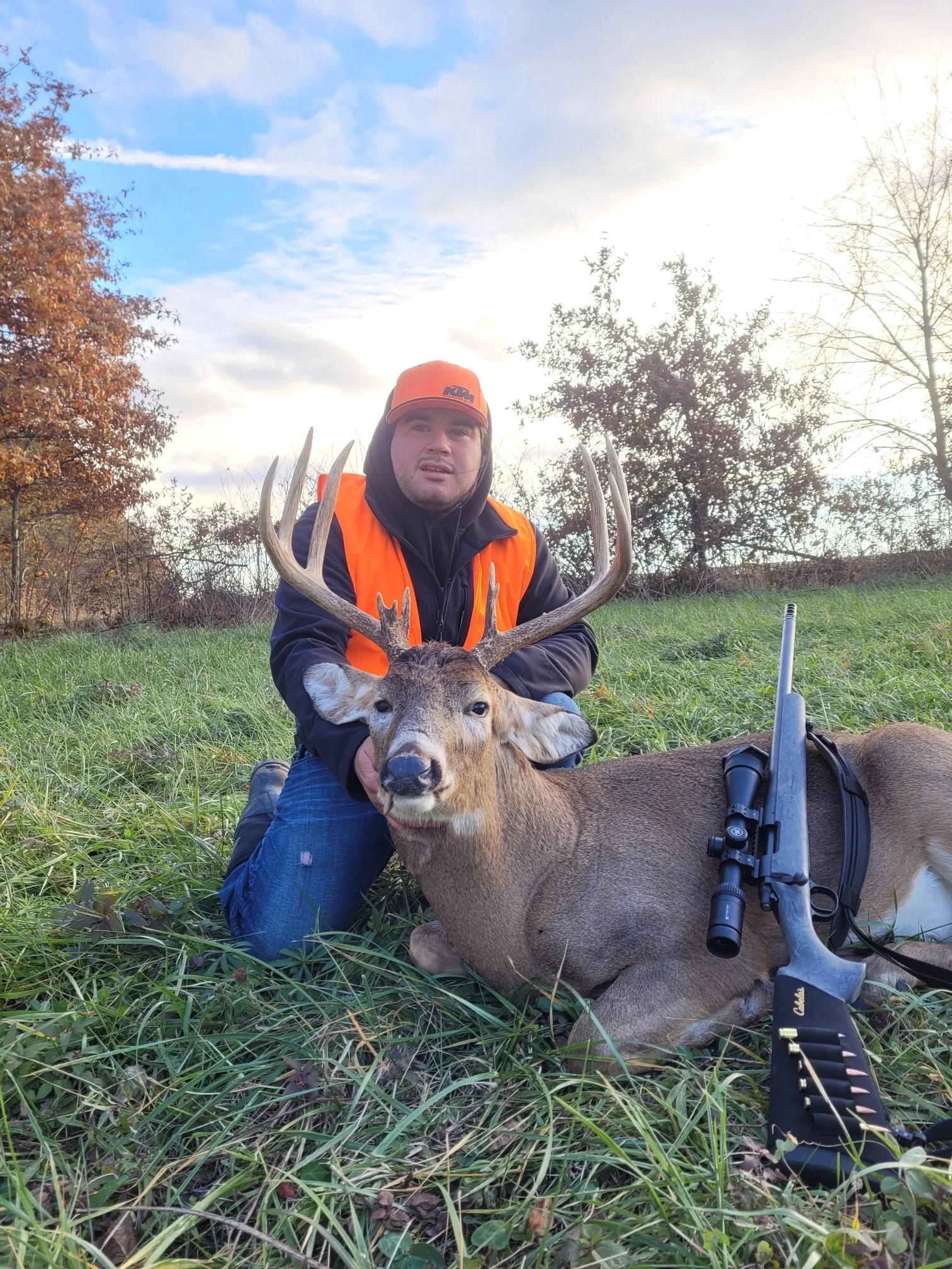 A man kneeling in a grassy field holding a large deer with antlers. The man is wearing an orange hunting vest and a cap, and there is a rifle lying on the ground beside the deer. Trees with autumn leaves are in the background under a partly cloudy sky.