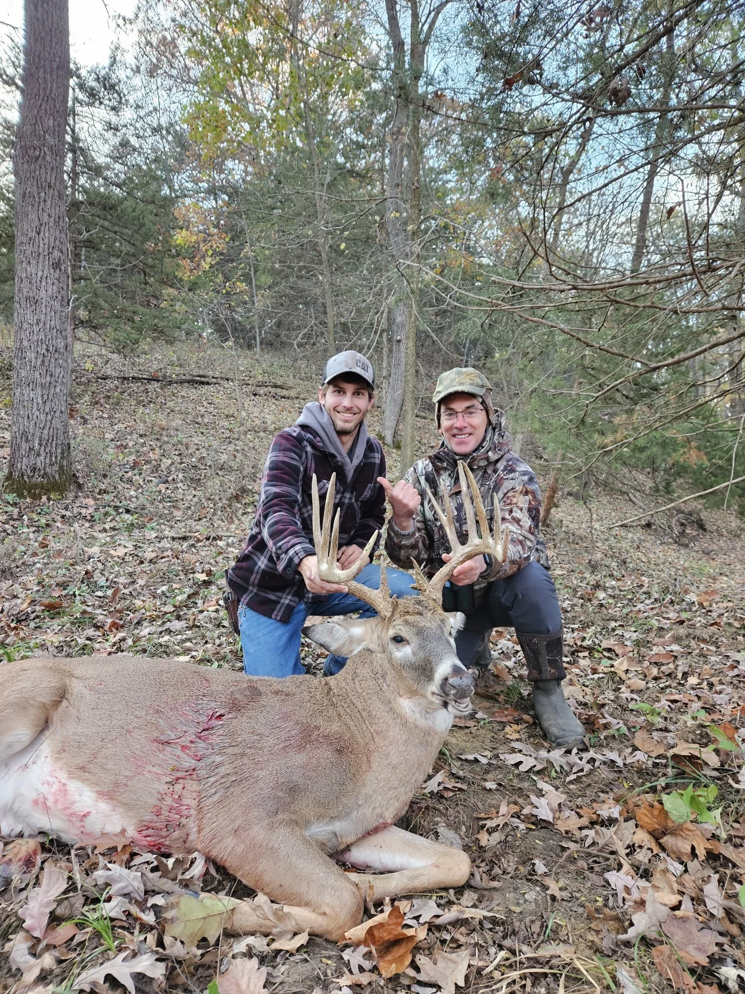 Two men, one in a plaid jacket and gray hoodie and the other in camouflage, kneeling in a wooded area with fallen leaves, holding antlers of a freshly hunted deer with a wound on its side, while smiling at the camera.