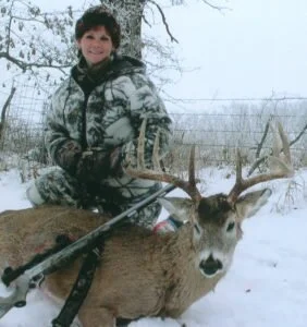 A young boy in winter clothing kneels beside a dead deer with antlers, holding a rifle during winter.