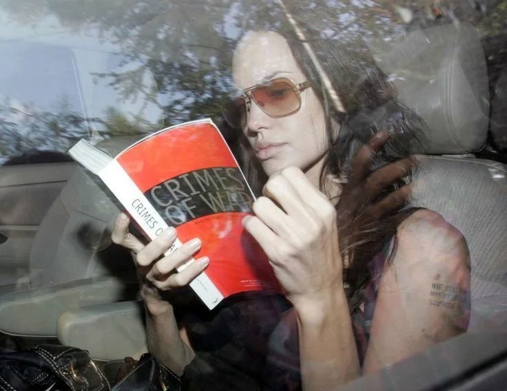 Angelina Jolie wearing sunglasses while reading Crimes of War inside a car.