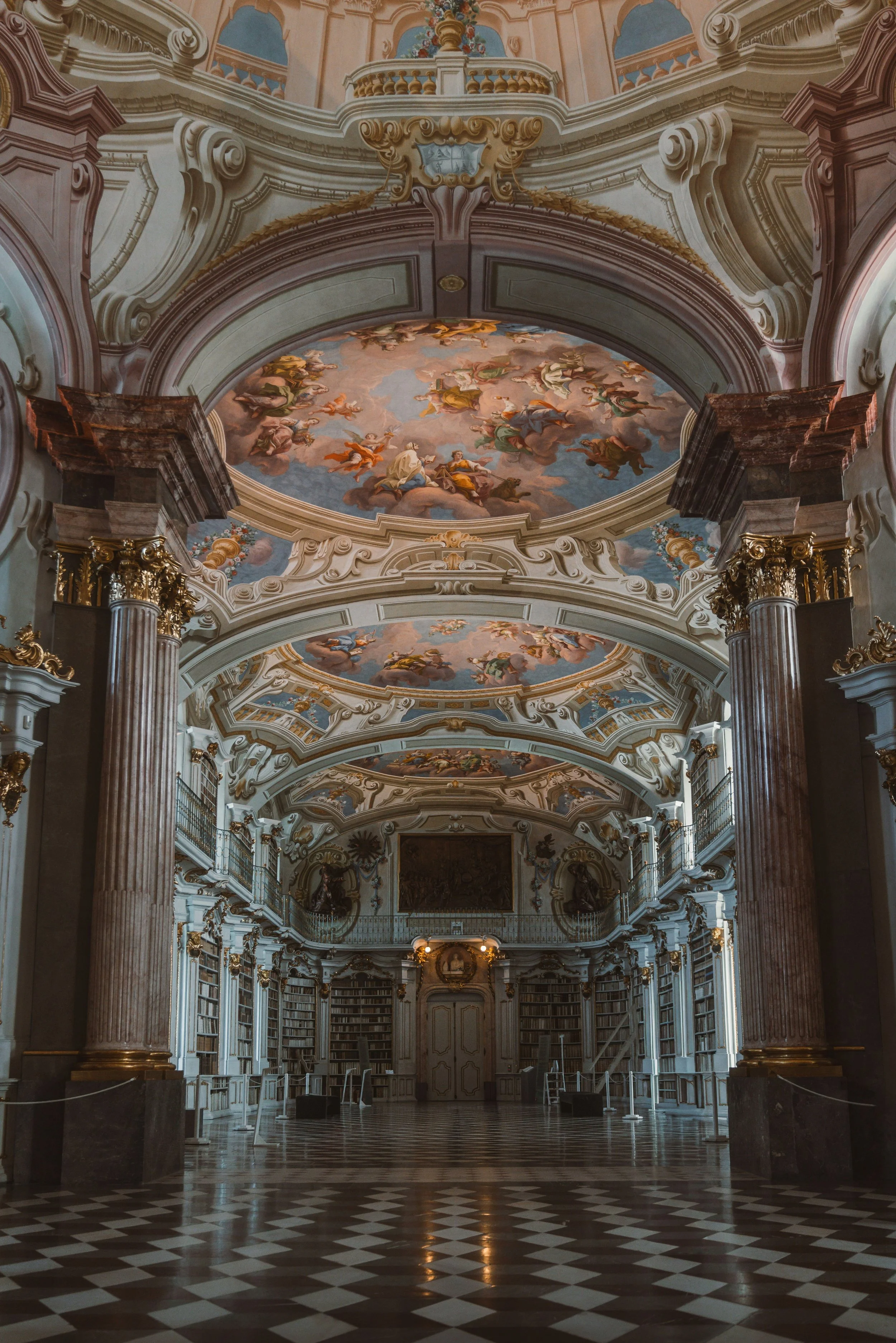 Library with ornate baroque hall with frescoed ceilings, marble columns, and a patterned stone floor.