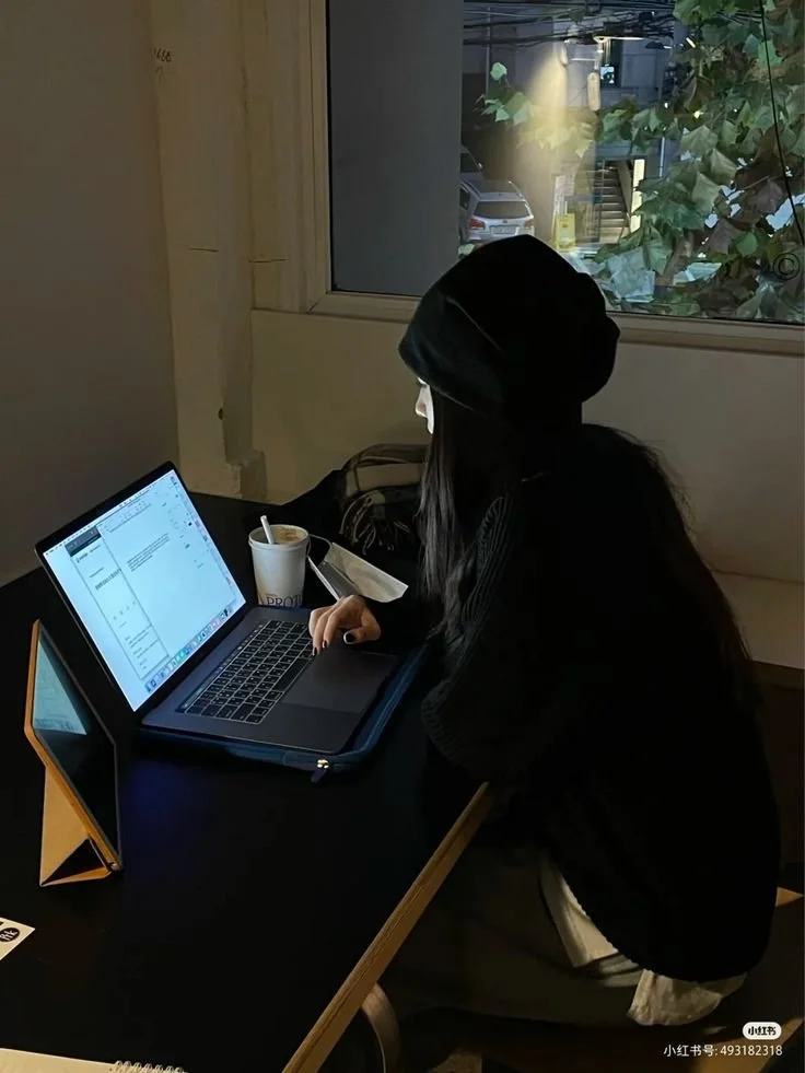 Girl in a black sweater with black hat working on a laptop at a small table with a coffee cup beside her.