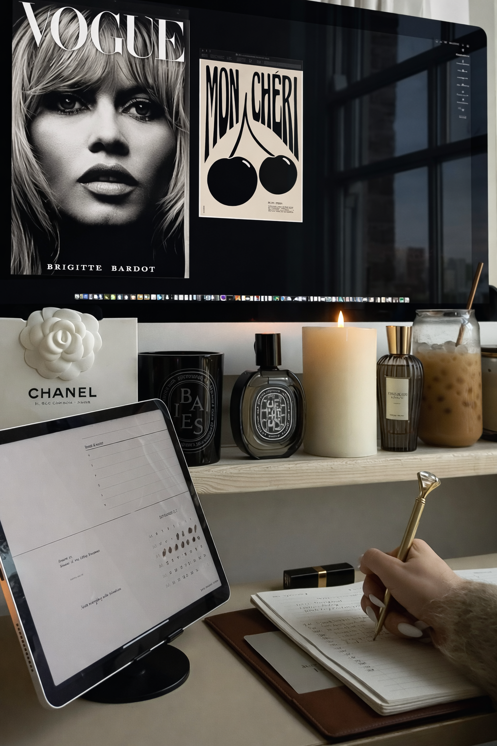 Elegant writing workspace with a desktop monitor, luxury candles and perfume, iced coffee, a tablet planner, and a hand writing in a notebook—styled with a chic, editorial aesthetic for a self-publishing website.