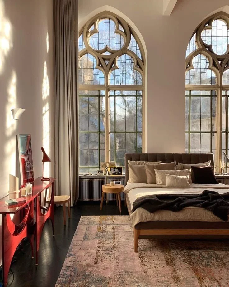 Sunlit bedroom with tall arched windows, a large bed with neutral bedding, and a red console table along the wall.